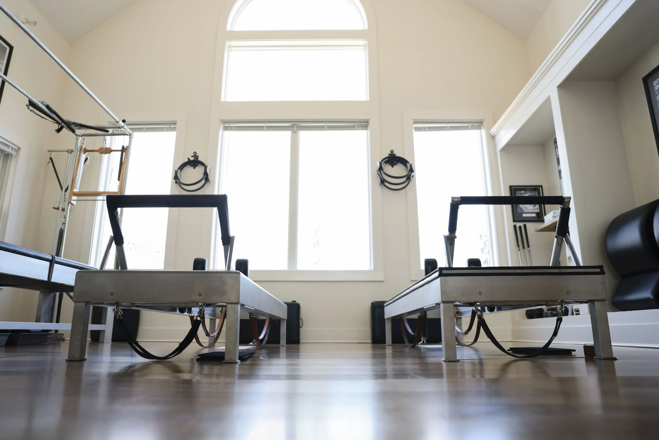 Pilates reformer machines in a bright, spacious home gym with tall windows, light-colored walls, and wooden flooring.