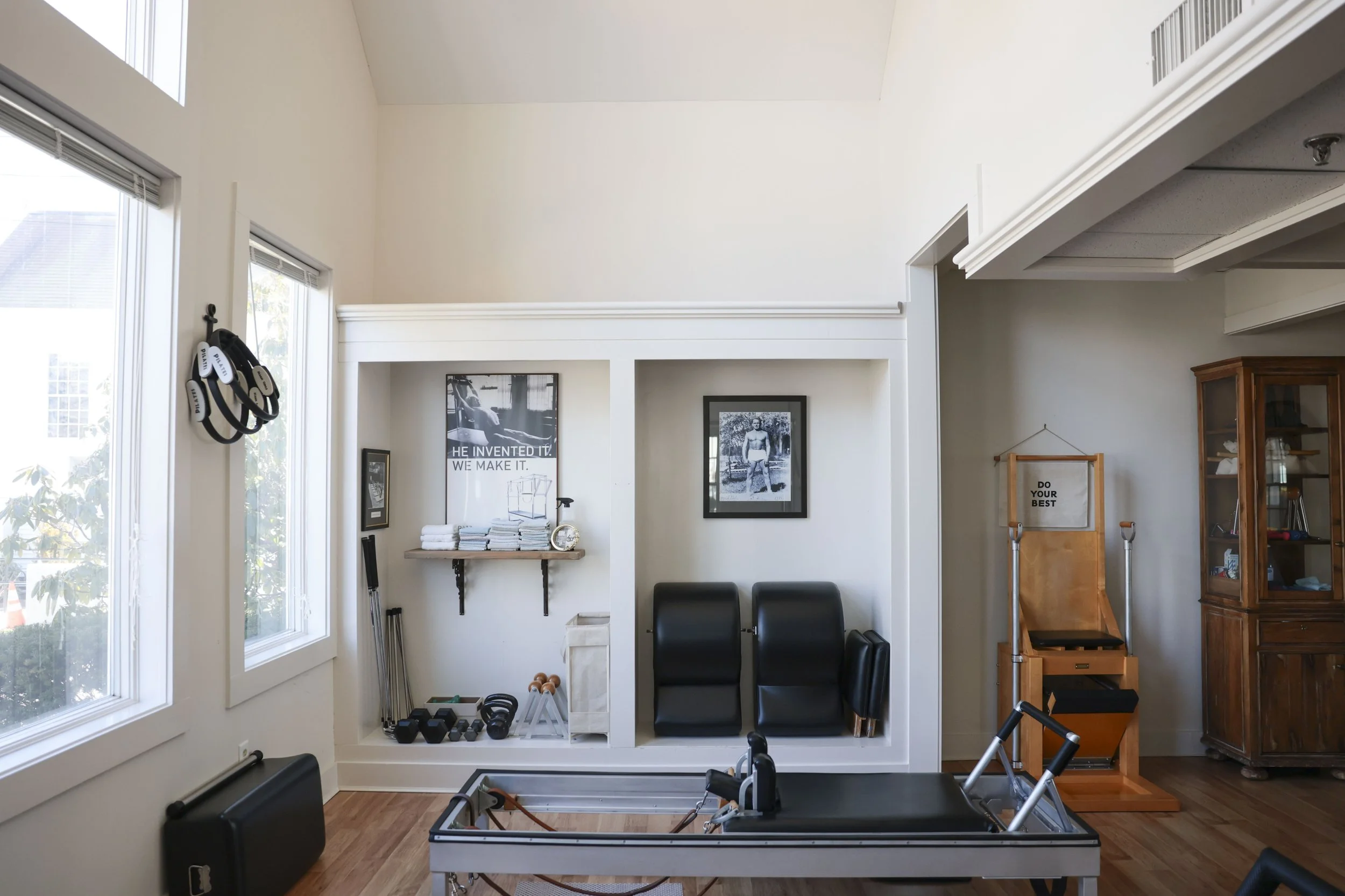 Home gym with windows, workout equipment, black chairs, framed pictures, and a wooden cabinet.