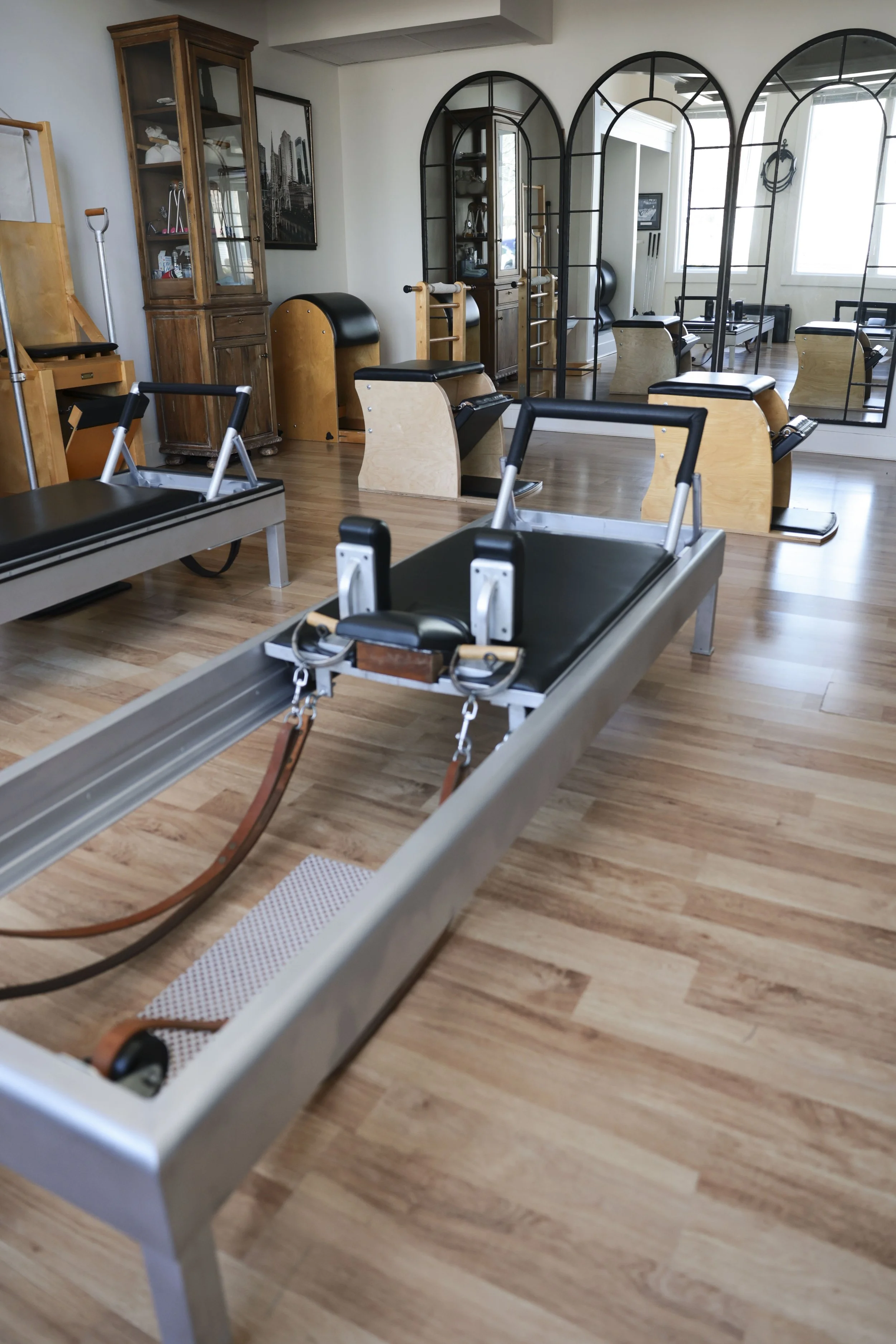 A Pilates studio with reformer machines, wooden chairs, a glass display cabinet, and large mirrors reflecting the room.