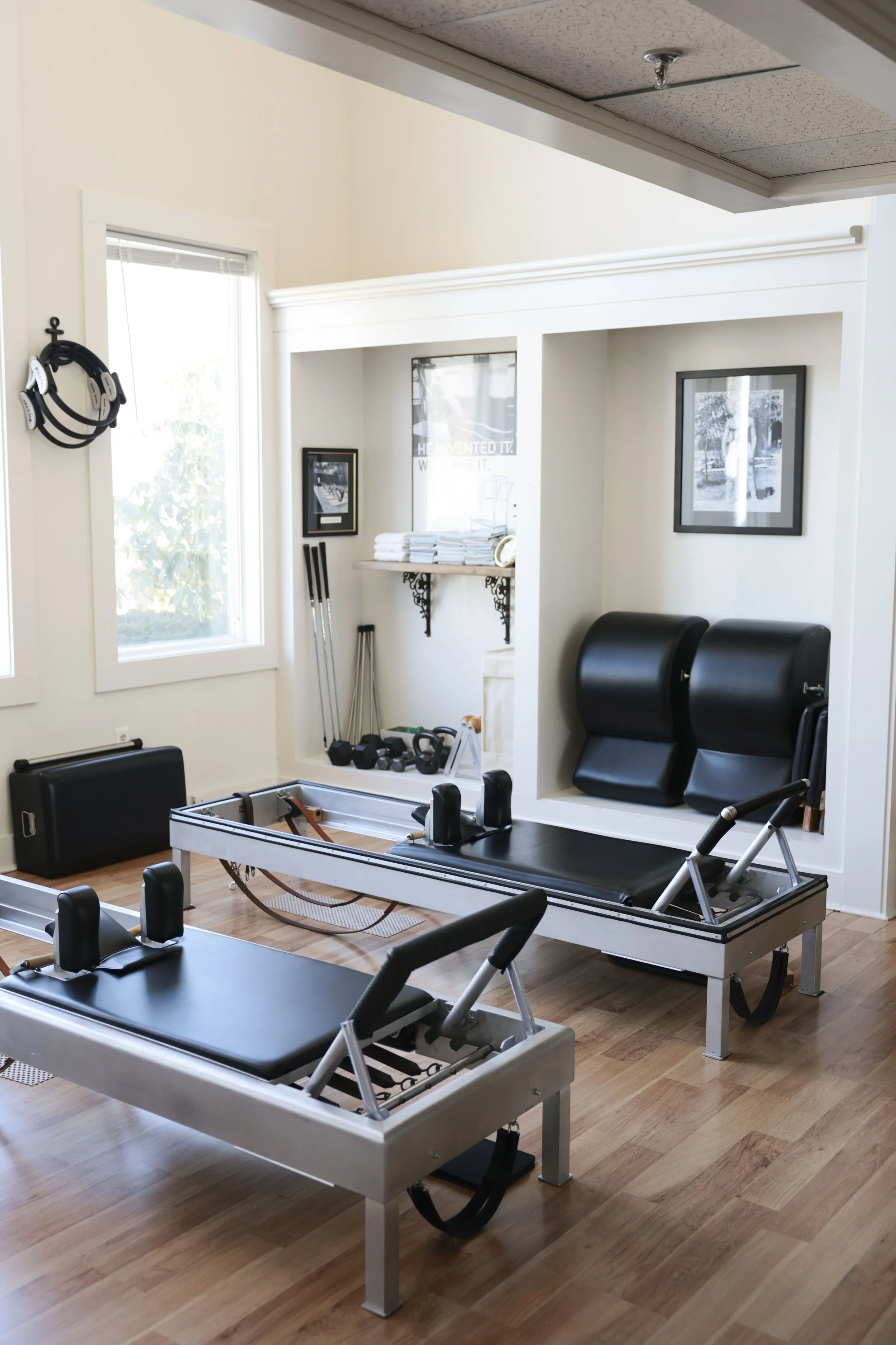 A professional physical therapy or rehabilitation clinic with specialized equipment including two reformer Pilates machines, a foam roller, dumbbells, resistance bands, and a shelf with towels. The room has windows, framed black-and-white photographs
