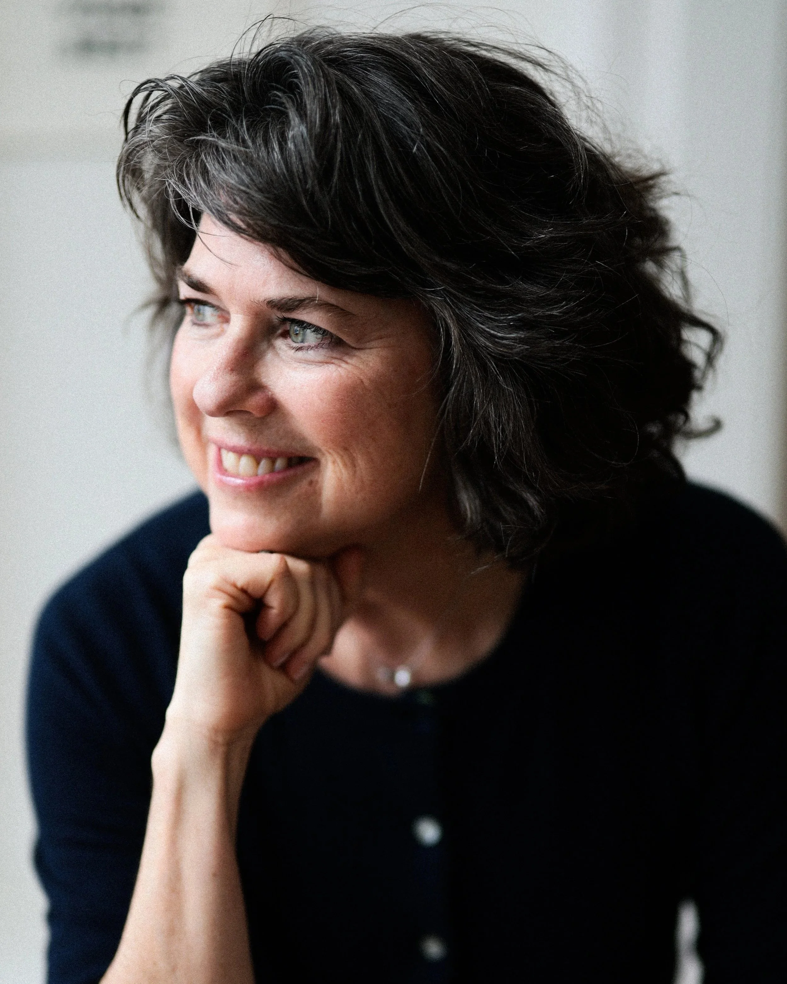 A woman with dark, wavy hair and light eyes, smiling, resting her chin on her hand, wearing a dark top.