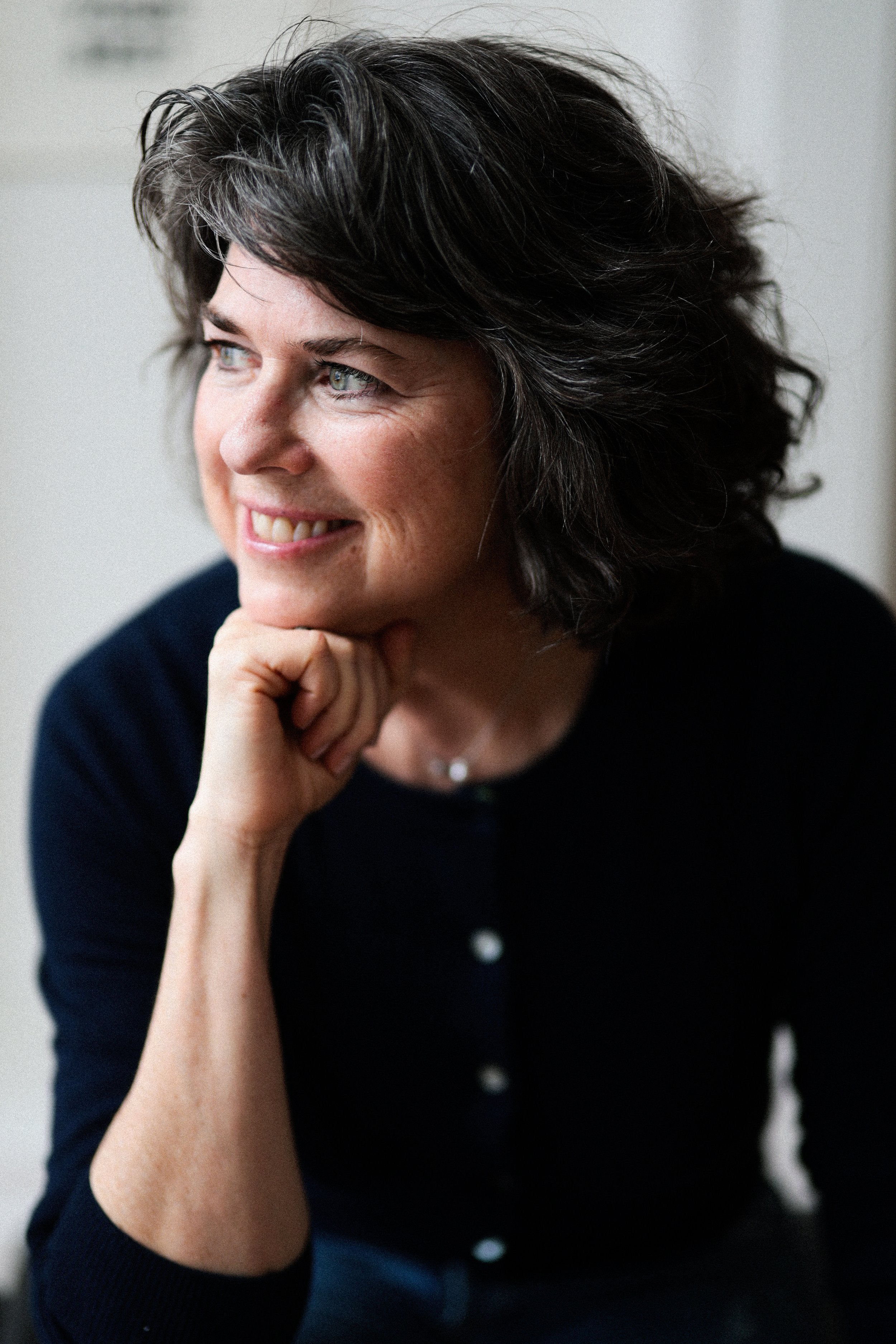 A woman with dark, wavy hair and fair skin smiling while resting her chin on her hand, looking to her right.