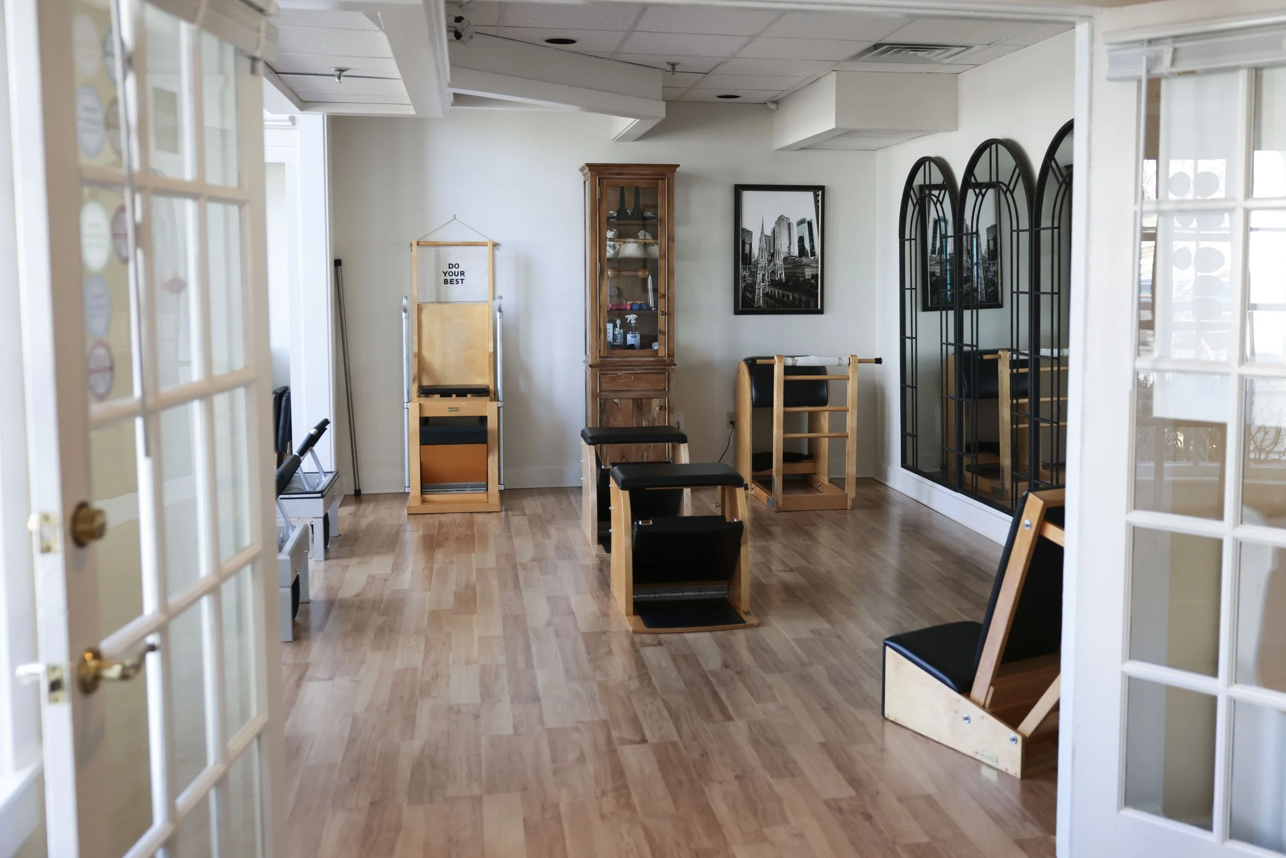 An interior room with wood flooring and white walls, containing Pilates equipment such as reformers and chairs, with framed photos on the wall and a wooden display cabinet.