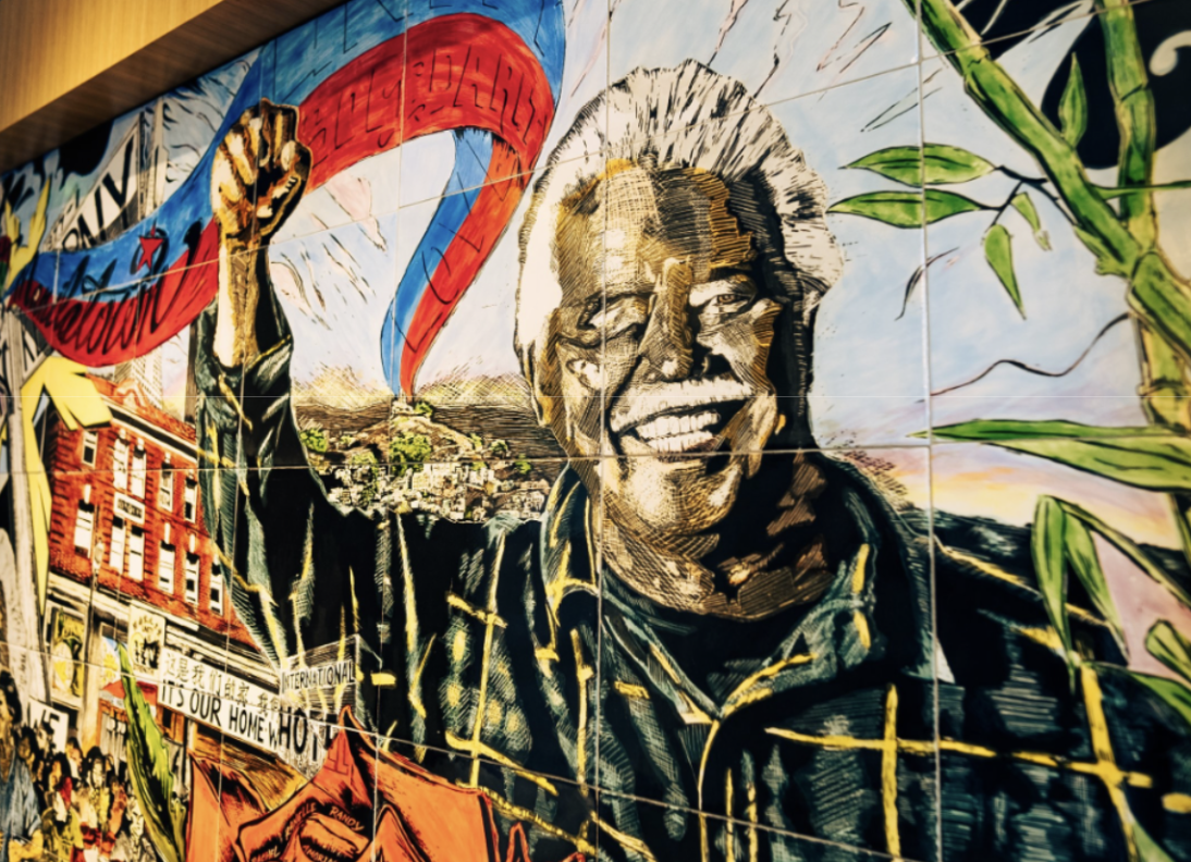 colorful mural of asian man with fist in air surrounded by buildings and greenery and a blue and red and white flag or banner
