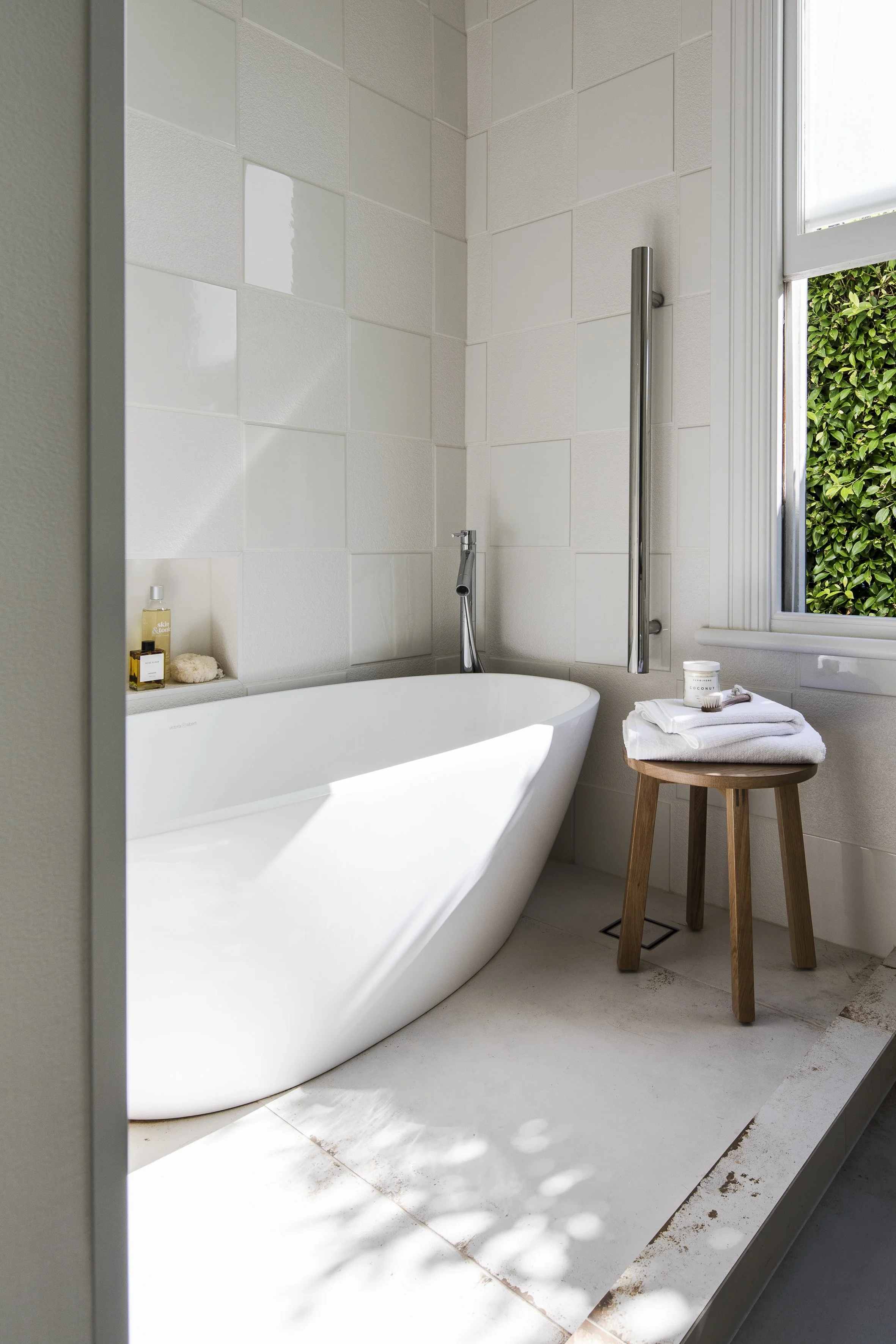 Modern bathroom renovation auckland with freestanding white bathtub, wooden stool with towels and candle, white tiles, and a window with green foliage outside.