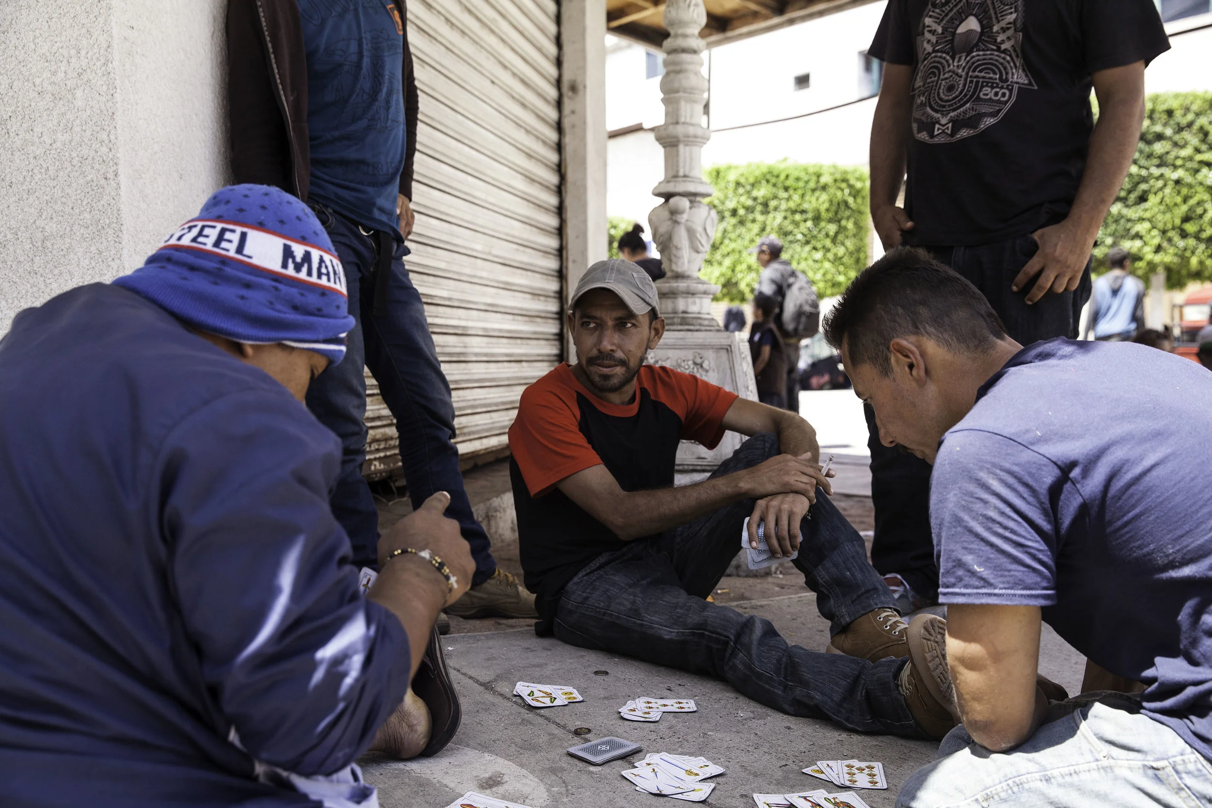 People wait outside under the shade to seek advice from attorneys near the U.S. - Mexico border in Tijuana, MX. Carlos (center) from Honduras plays cards to pass the time with other people who have traveled from Central America to seek asylum in the United States on April 28, 2018.
