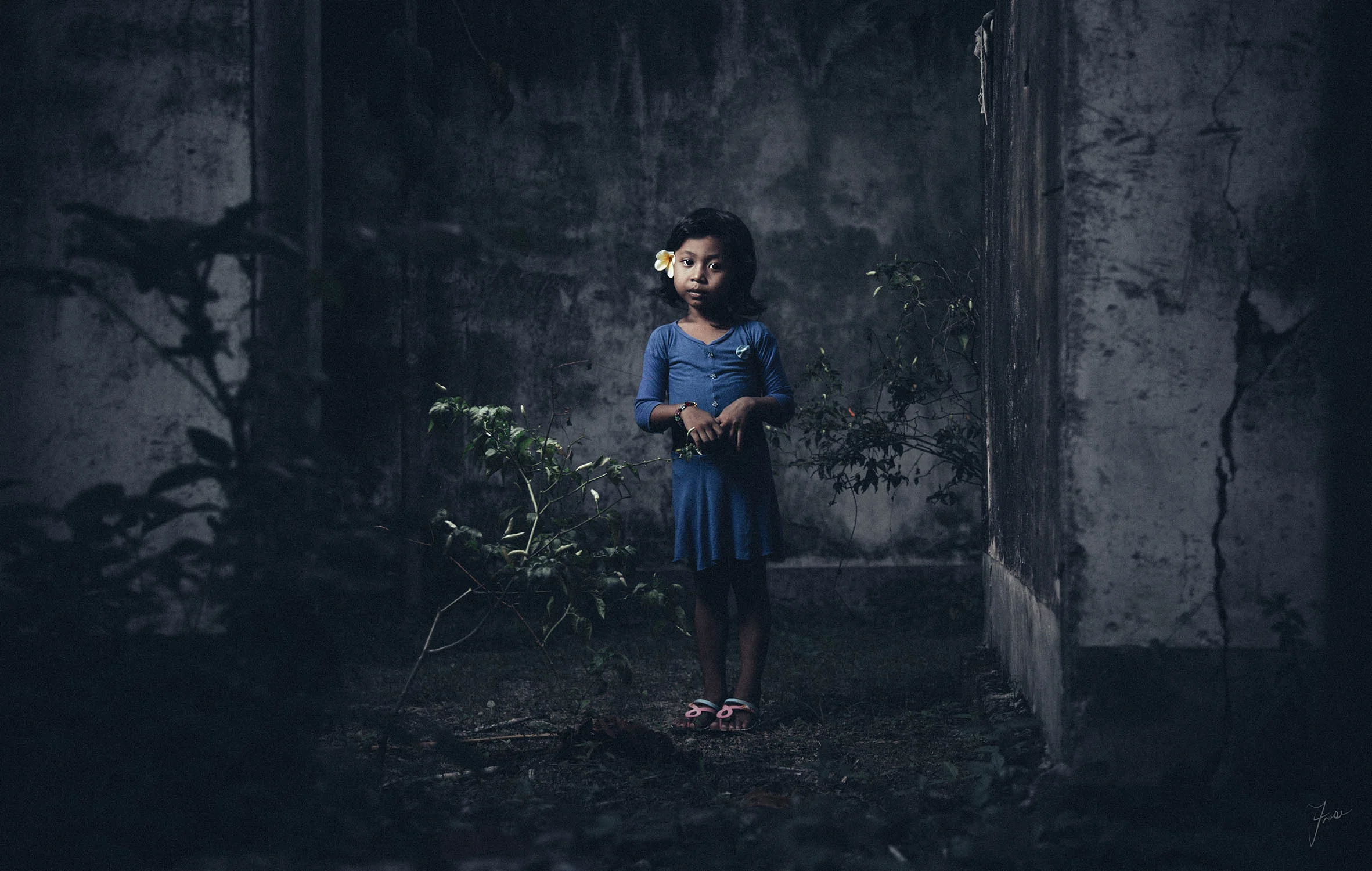Dramatic fine art portrait of a young girl in a blue dress with a yellow frangipani flower in her hair, standing among overgrown plants inside a weathered abandoned building, lit with cinematic blue tones — photography by Fraser