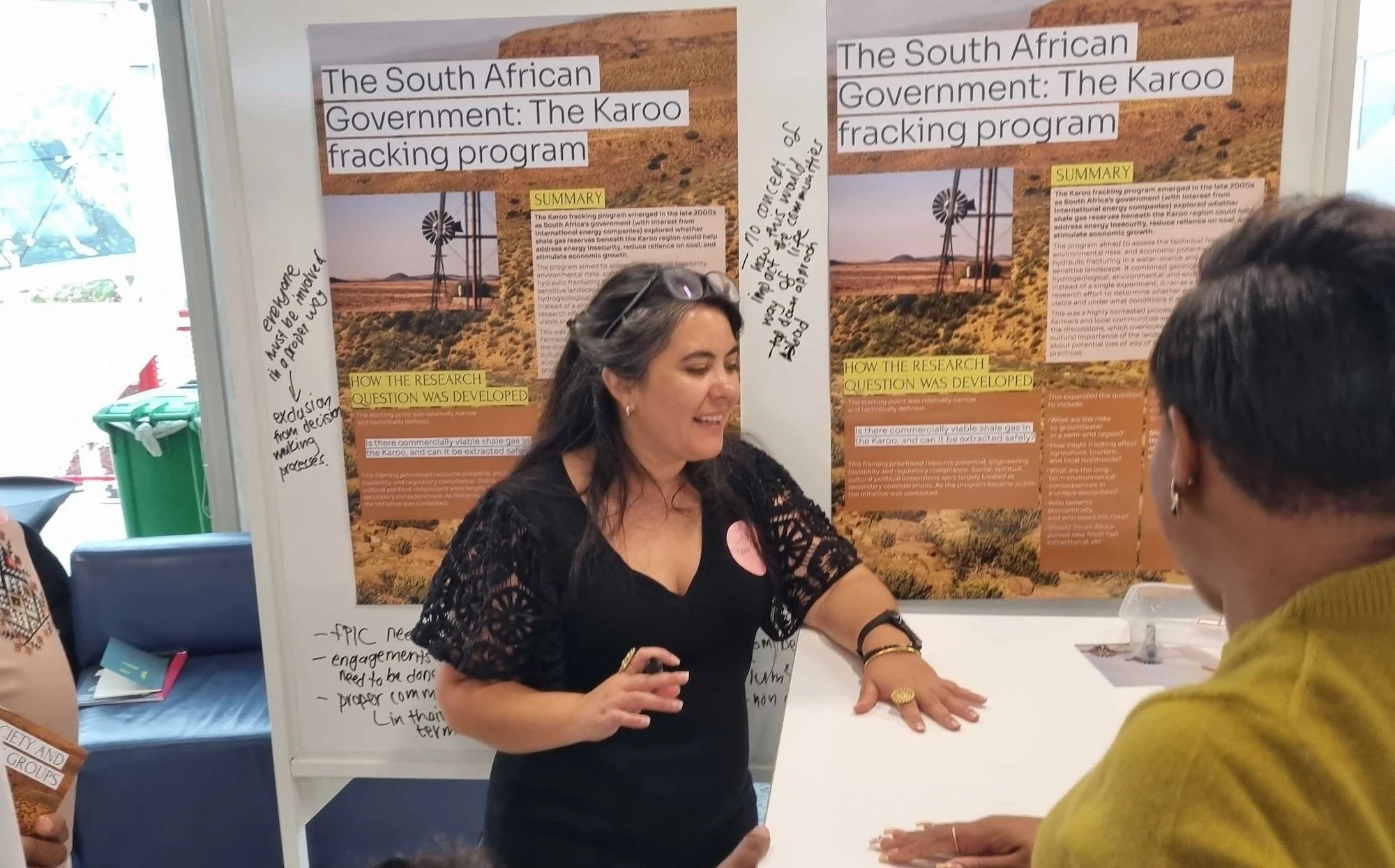 a person stands in front of a whiteboard covered with pictures and writing, showing something on a table to people who are standing around