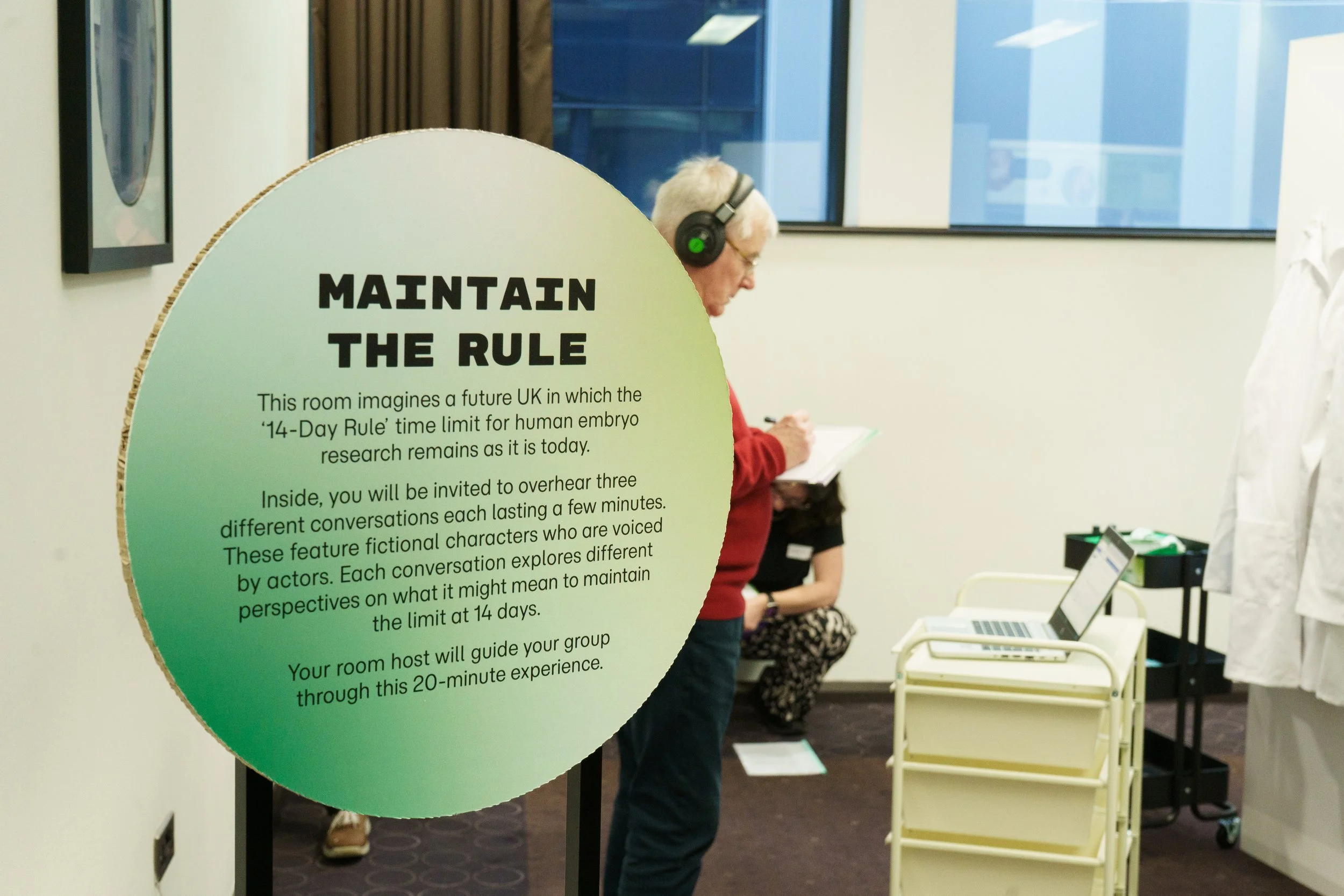 A person with headphones on, writing on a notepad stands behind a large colourful circular poster entitled 'Maintain the Rule'