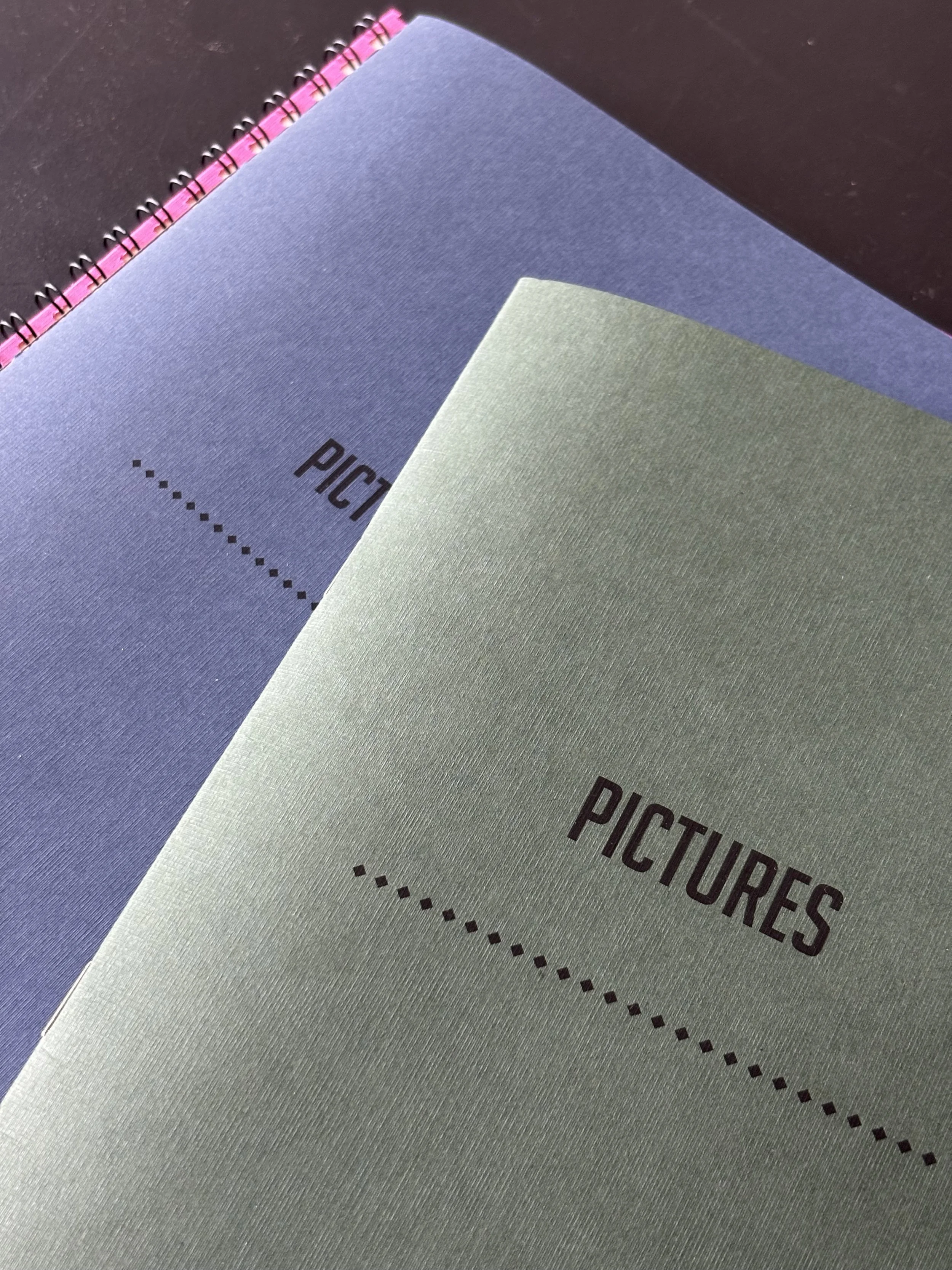 Close-up of three notebooks stacked, with the top one labeled 'PICTURES' in capital letters, on a dark surface.