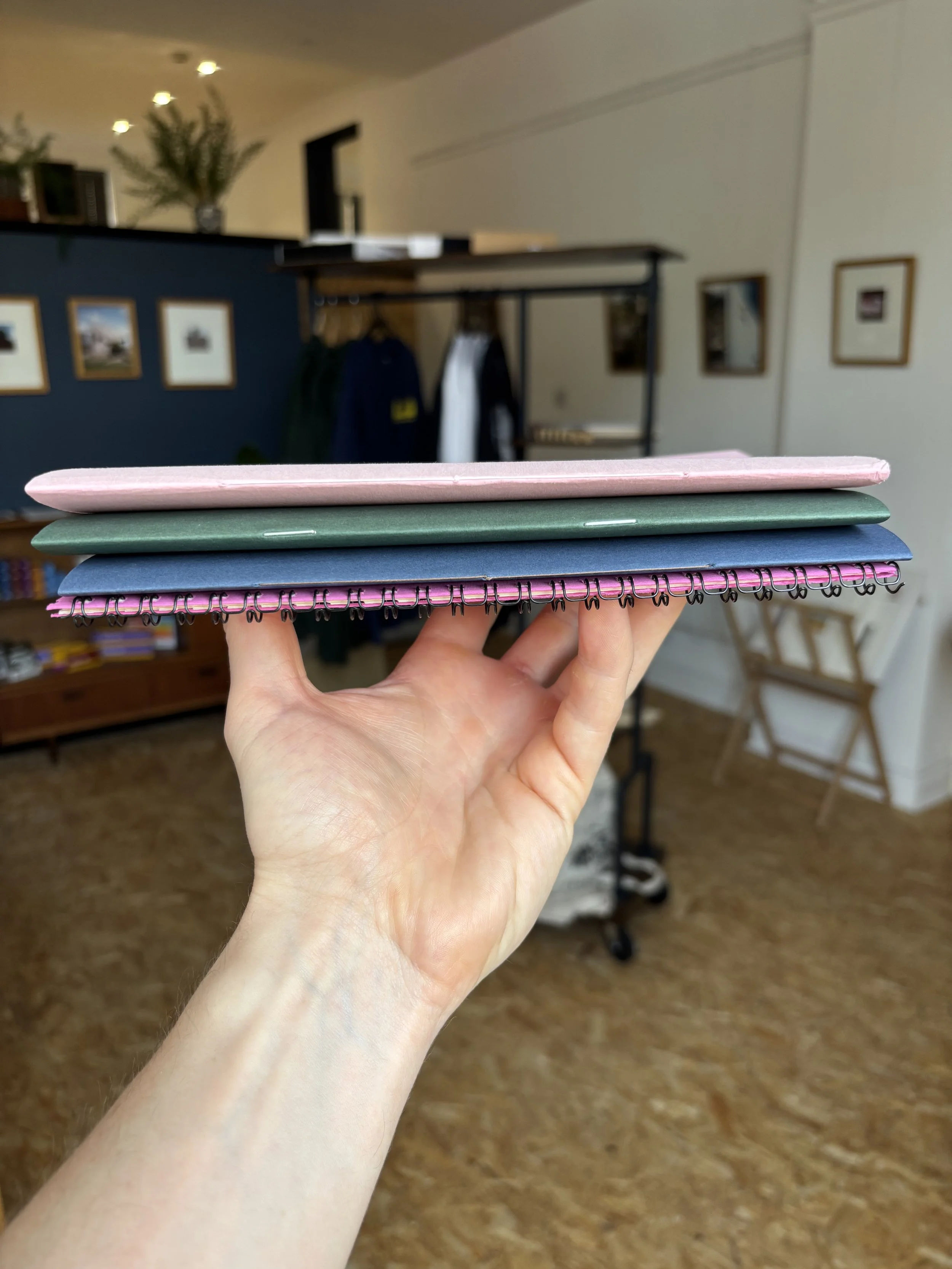 Hand holding three small notebooks or notepads in pink, green, and blue colors, with a blurred background of a room showing framed pictures on the walls, a desk with clothes hanging, a bookshelf, and a wooden chair.