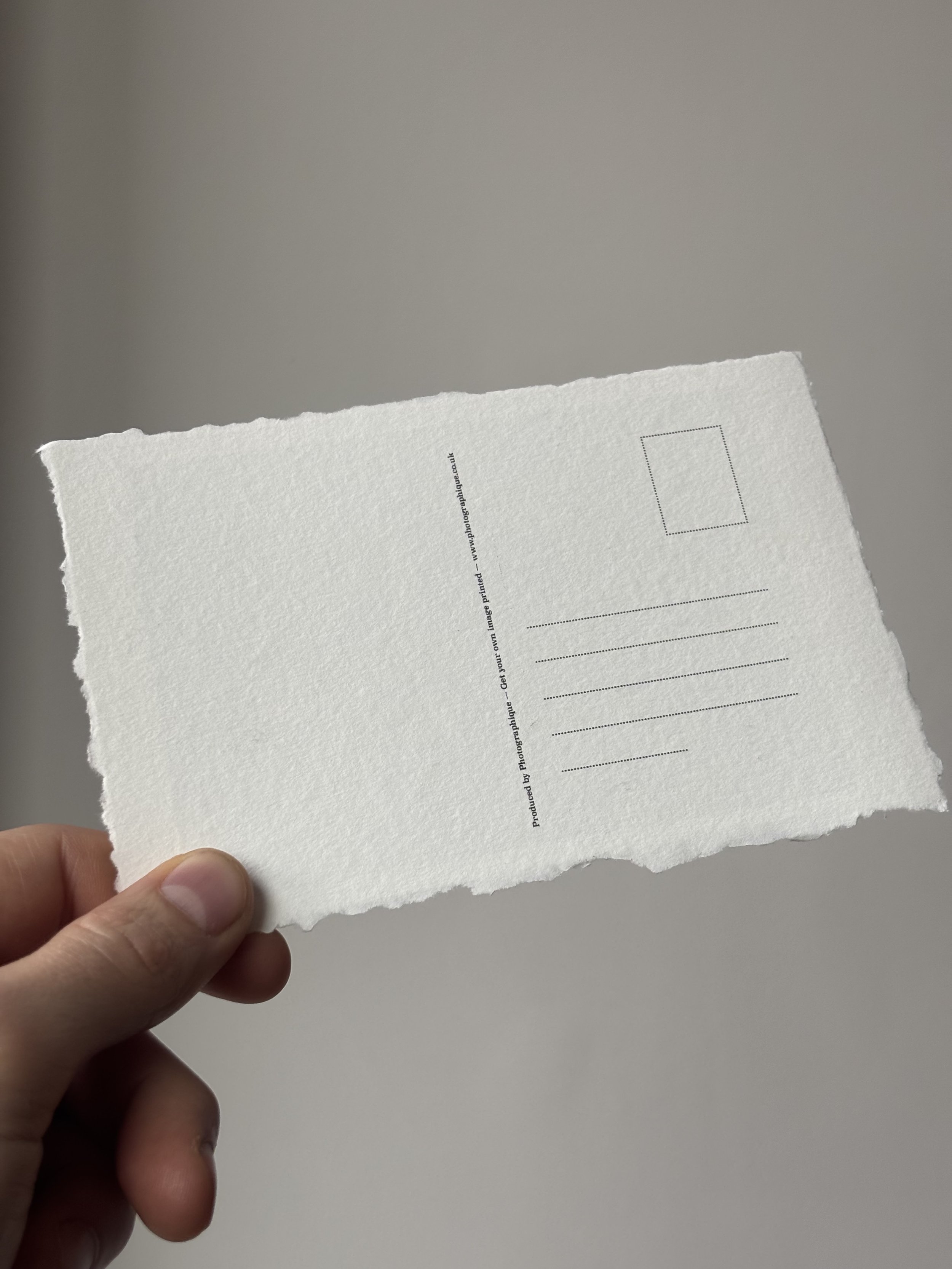 Person holding a white, textured postcard with black printed text and a dotted outline for a stamp, against a plain light gray background.