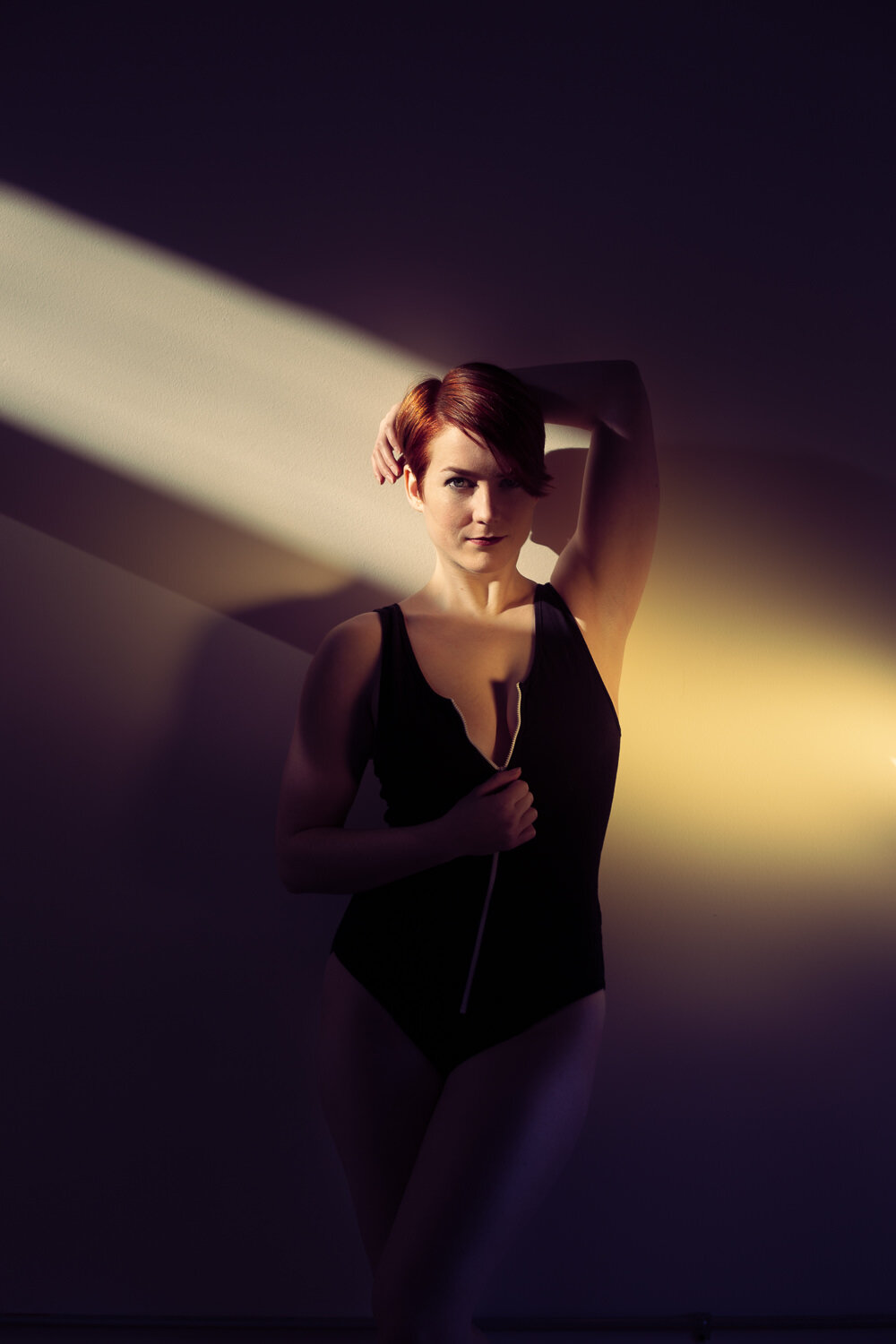 woman in black bodysuit and red hair leaning against wall in diagonal shaft of light