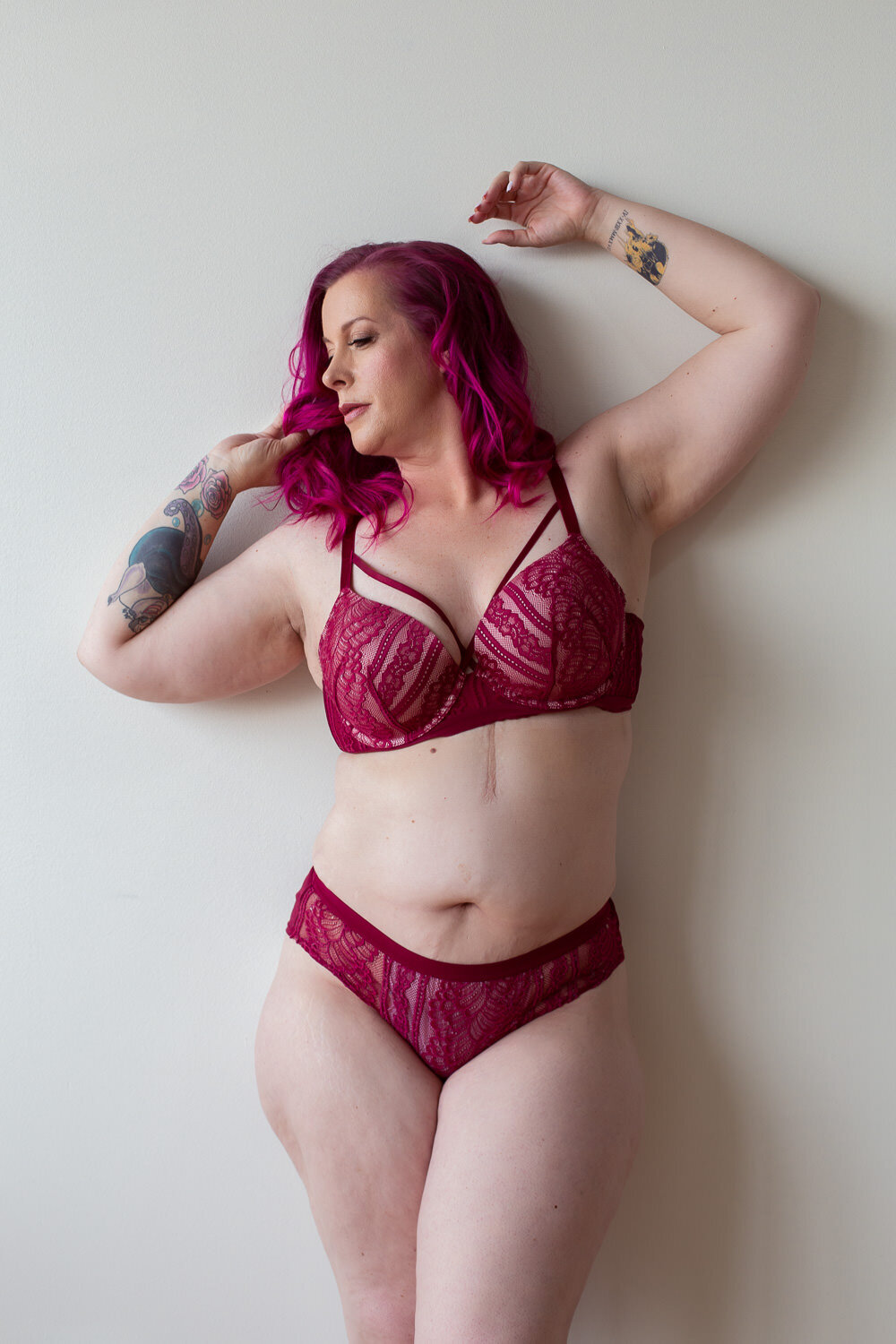 plus size woman with pink hair and matching pink bra and panty set leaning against a white wall