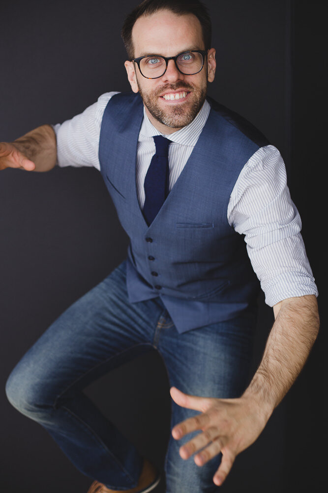 Man with dark hair glasses and beard wearing a blue vest and tie balancing on one foot