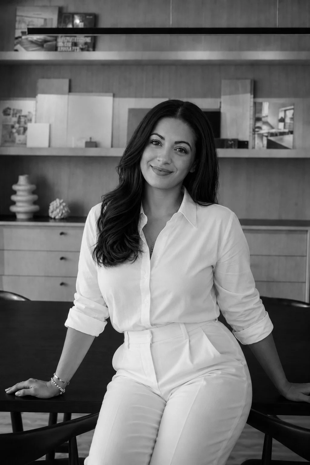 A woman with long dark hair, wearing a white shirt and white pants, sitting on a table in a modern indoor space with wooden shelves and decor in the background.