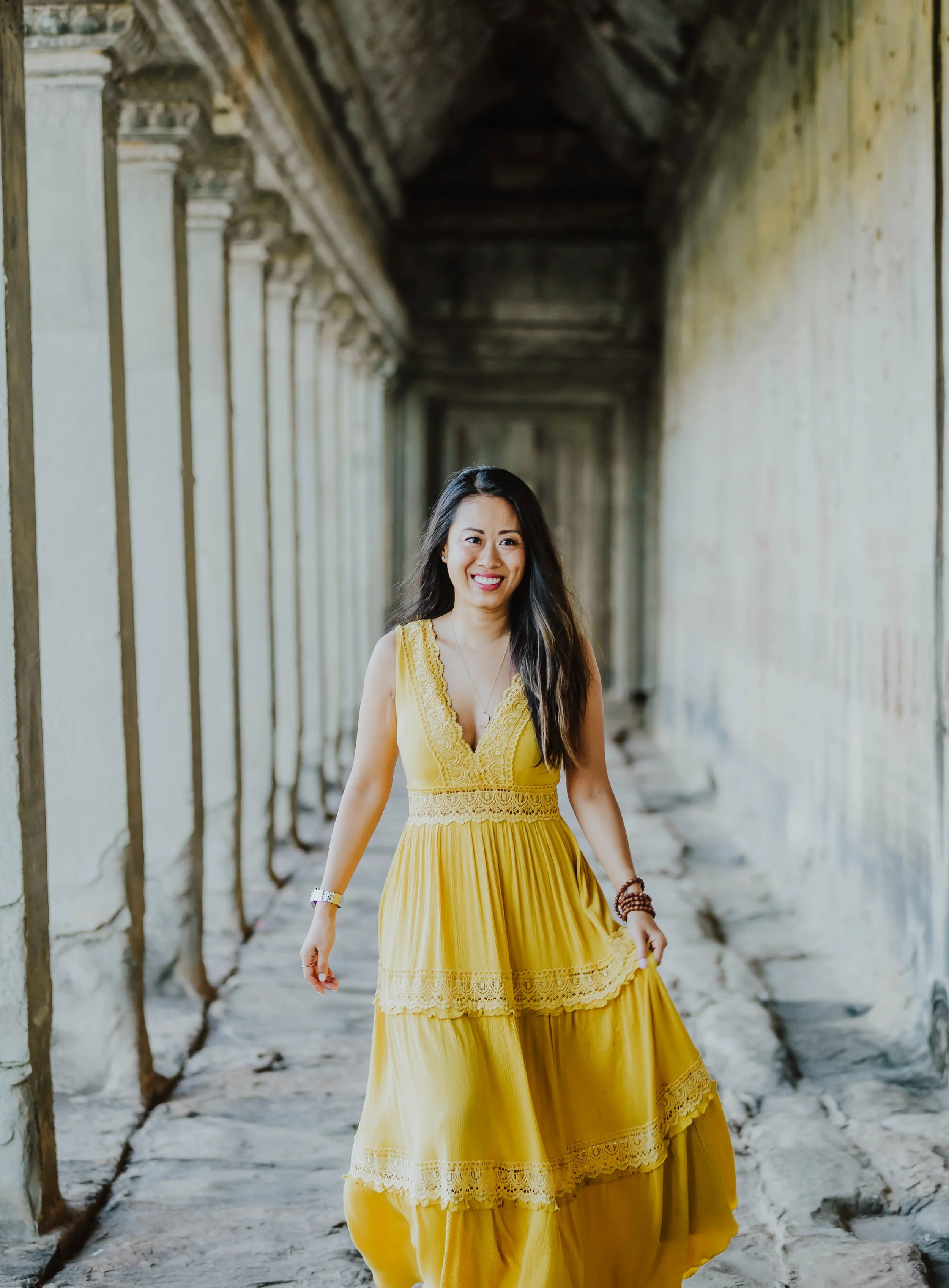 A woman with long dark hair smiling and wearing a yellow dress with lace details while walking through an ancient stone corridor with columns.