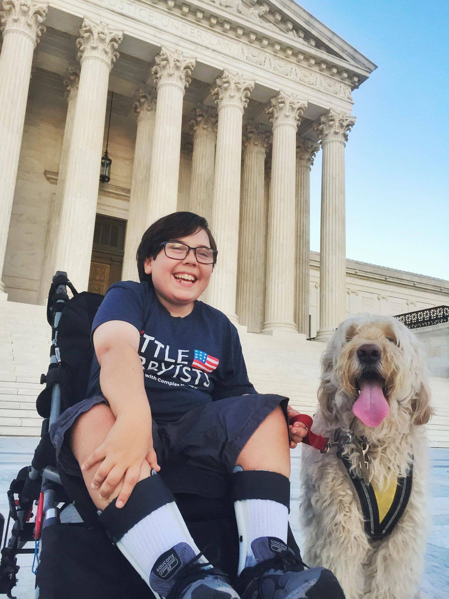  A young man wearing a Little Lobbyists t-shirt sits in his wheelchair, posing with his dog in front of the U.S. Supreme Court. 