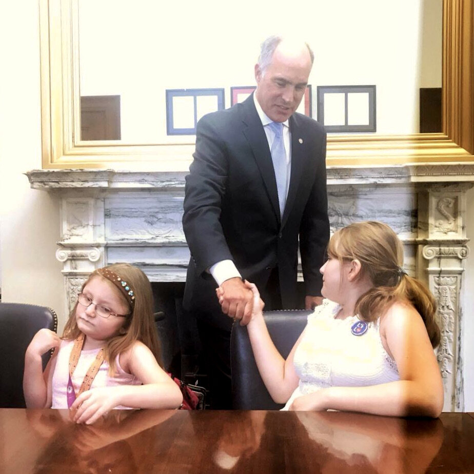  Two young girls are seated at a conference table in front of a marble fireplace. One shakes hands with her Senator. 