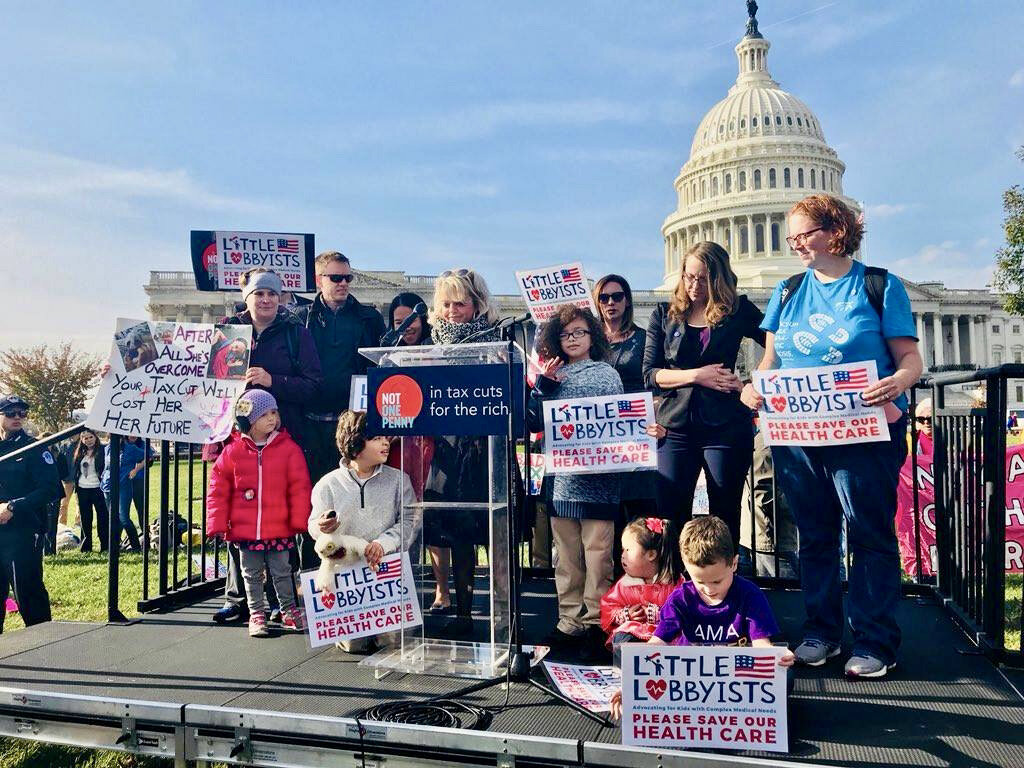  With the U.S. Capitol in the background, a group of speakers stands on a platform at a podium, delivering remarks on health care. 