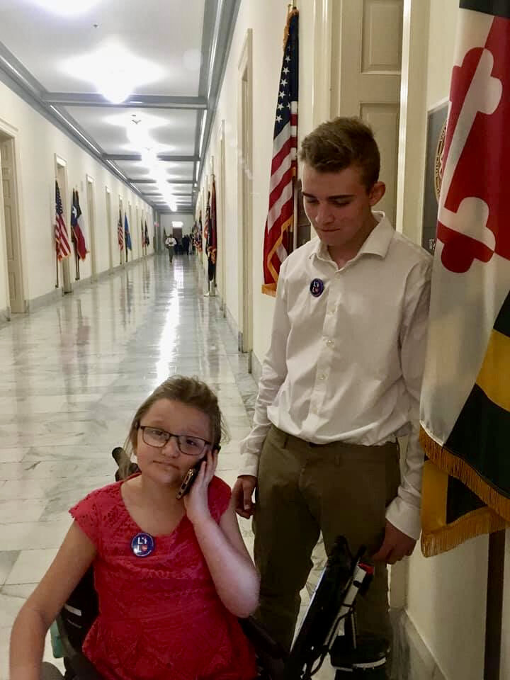  A young girl sits in her wheelchair and makes a phone call while her brother stands next to her. They are in a hall in a congressional office building. 