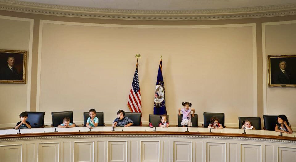  Several young children are seated at a long legislative dais, flanked by flags. 
