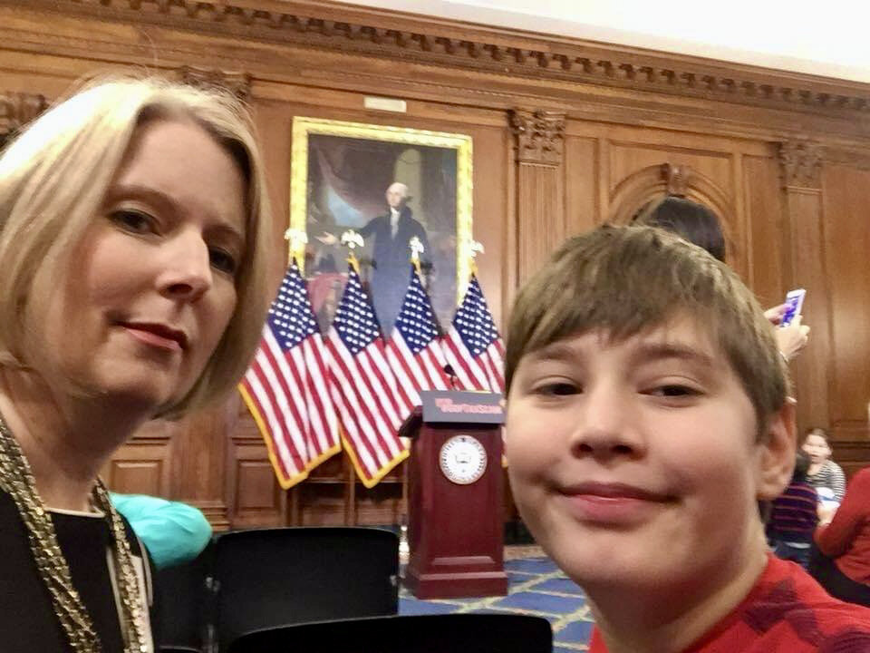  A mom and her son pose for a photo after a U.S. Senate press conference. The podium and U.S. flags are in the background. 