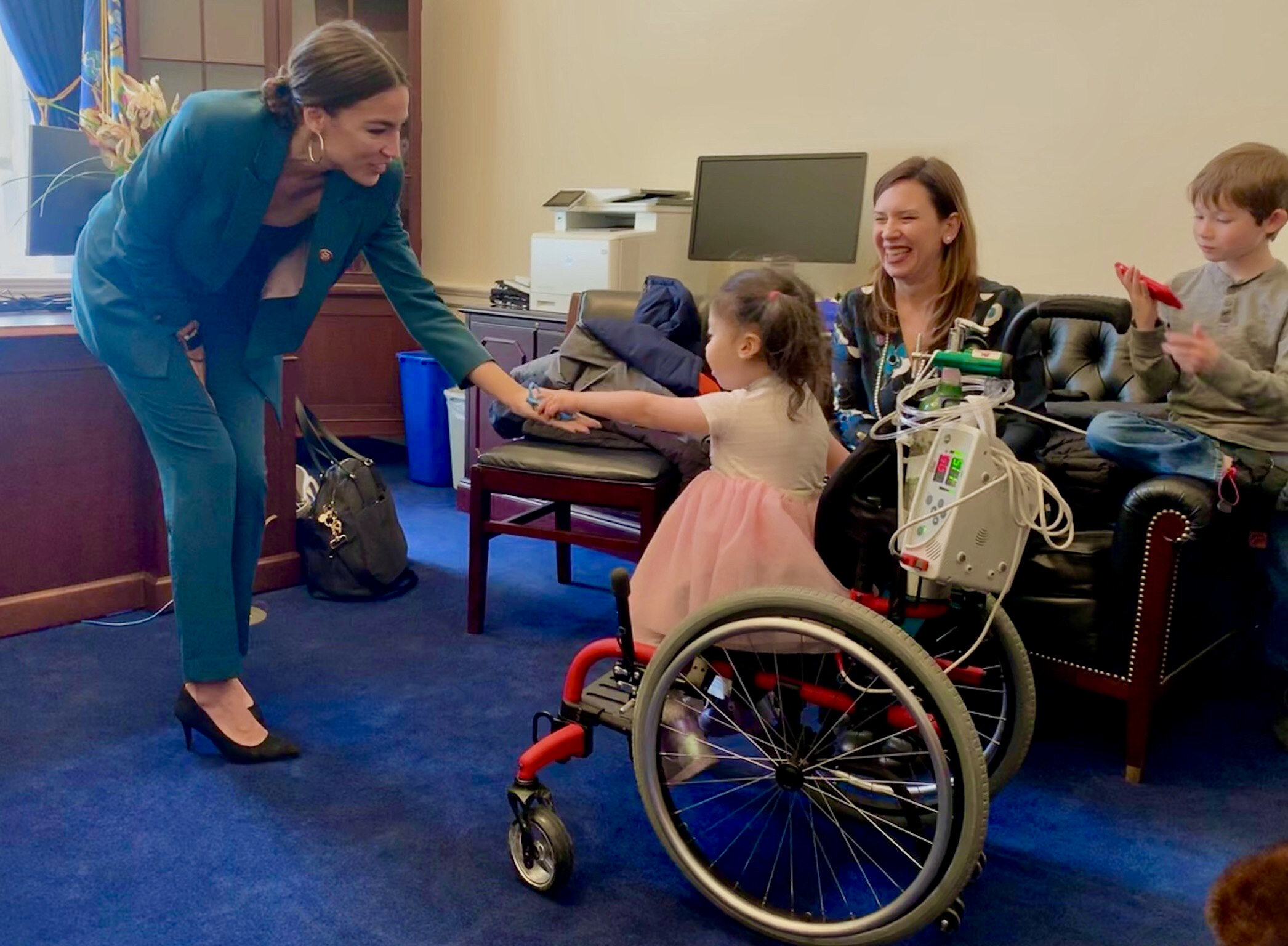  A new member of Congress wearing a vivid turquoise suit bends forward to grasp the hand of a Little Lobbyists child who reaches out to shake her hand. 