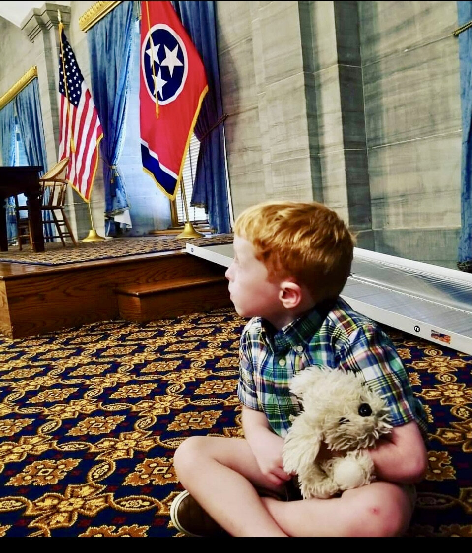 A young boy sits on a patterned carpet with state flags after a state legislative press conference. 