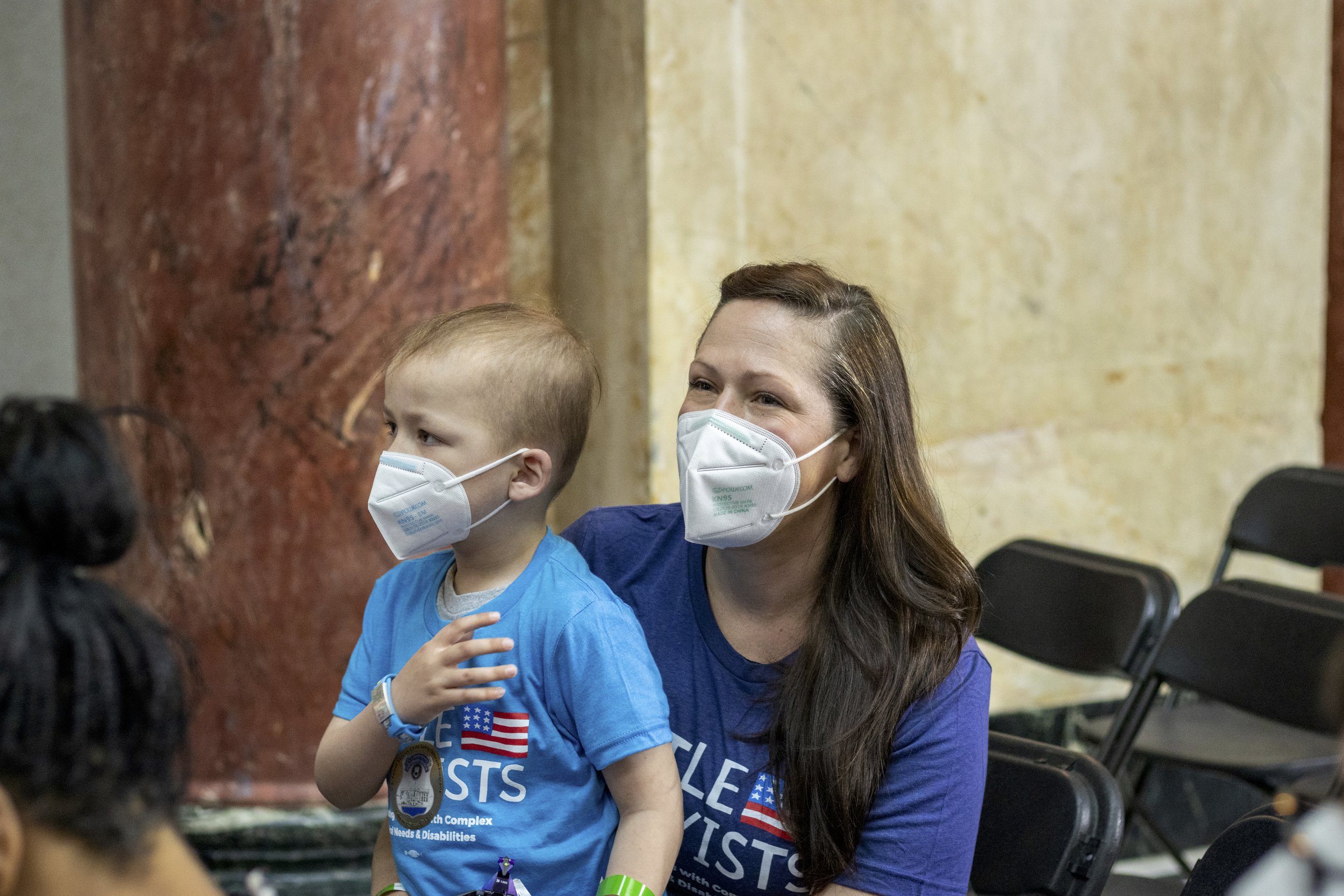  Megan holds her son Gabe in her lap inside the rally. Both are wearing N95 masks and they are seated alongside a beautiful marble wall in earth tones. 