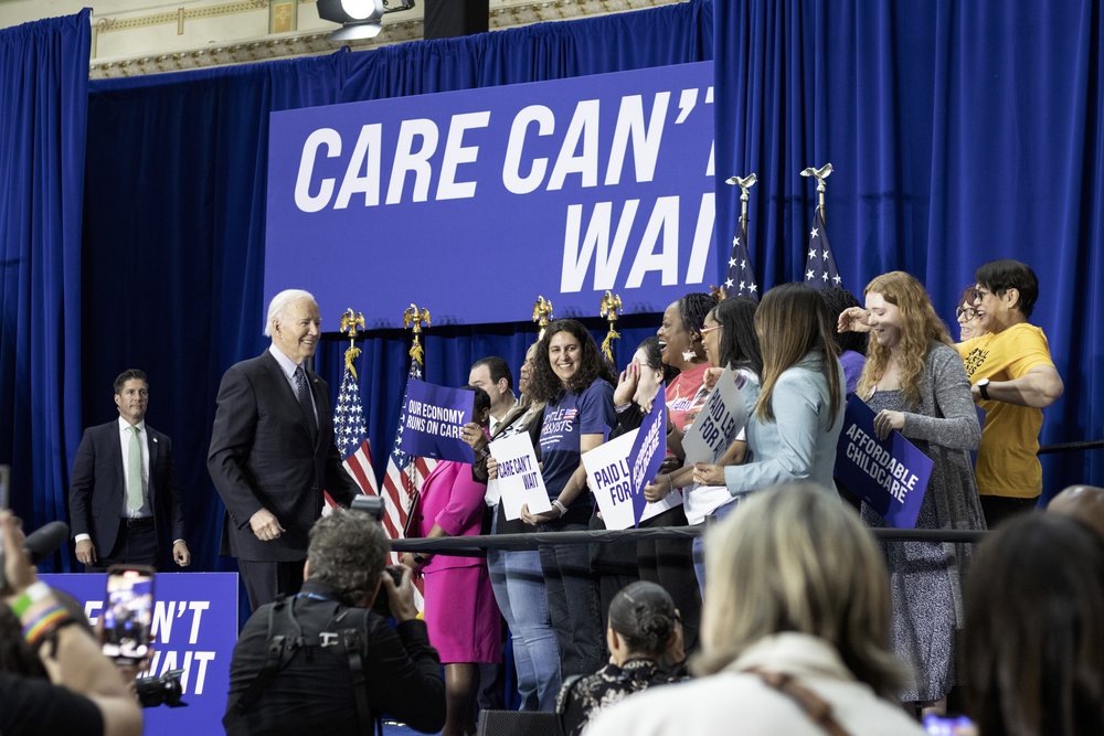  President Biden walks toward a group of caregivers to the right. They hold signs expressing support for care. Among them is Jamie, who has long, dark curly hair. Behind them is a large sign on the stage, royal blue with white lettering: Care Can’t W