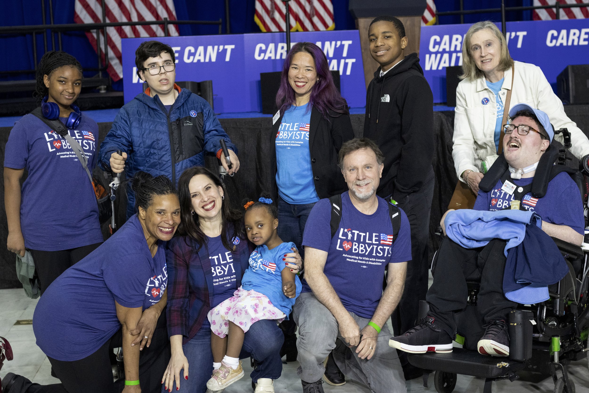  A group of us pose in front of the stage—some are using mobility devices, some are standing, some are kneeling on the floor.  