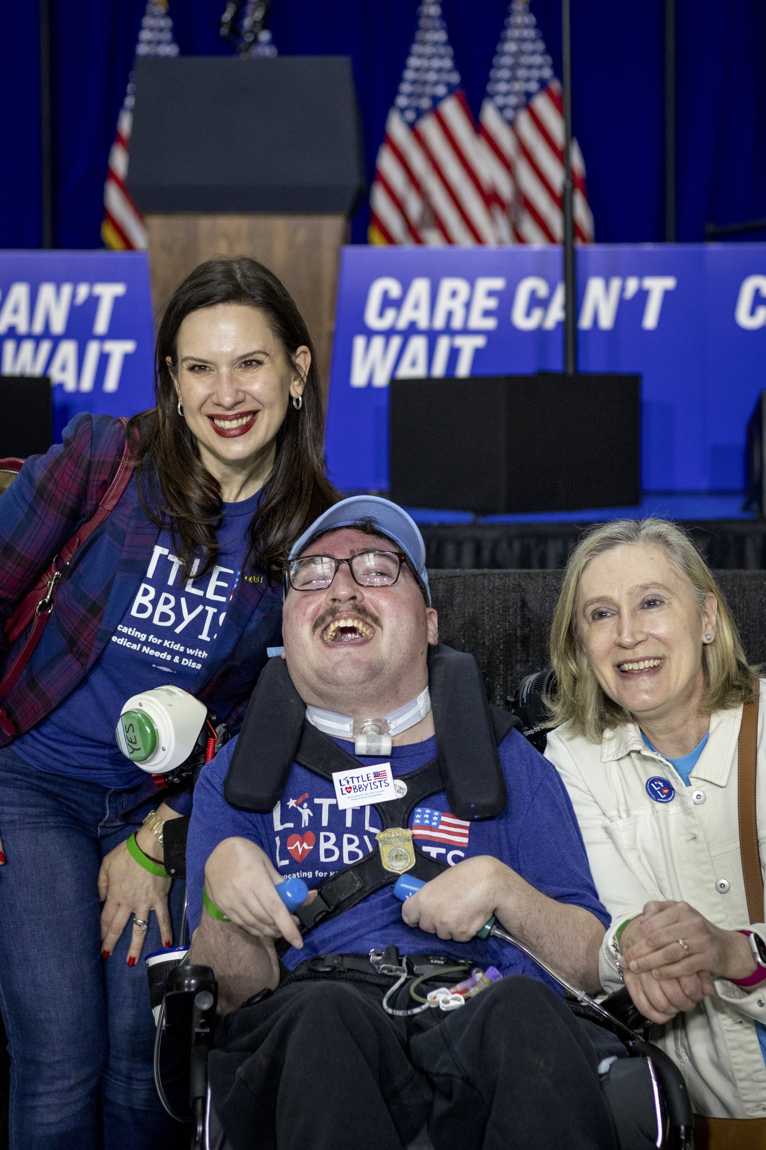  Laura and Jeneva pose on either side of Rob, who is smiling broadly from his wheelchair and wearing his LL baseball hat and his Clark-Kent-style glasses. 