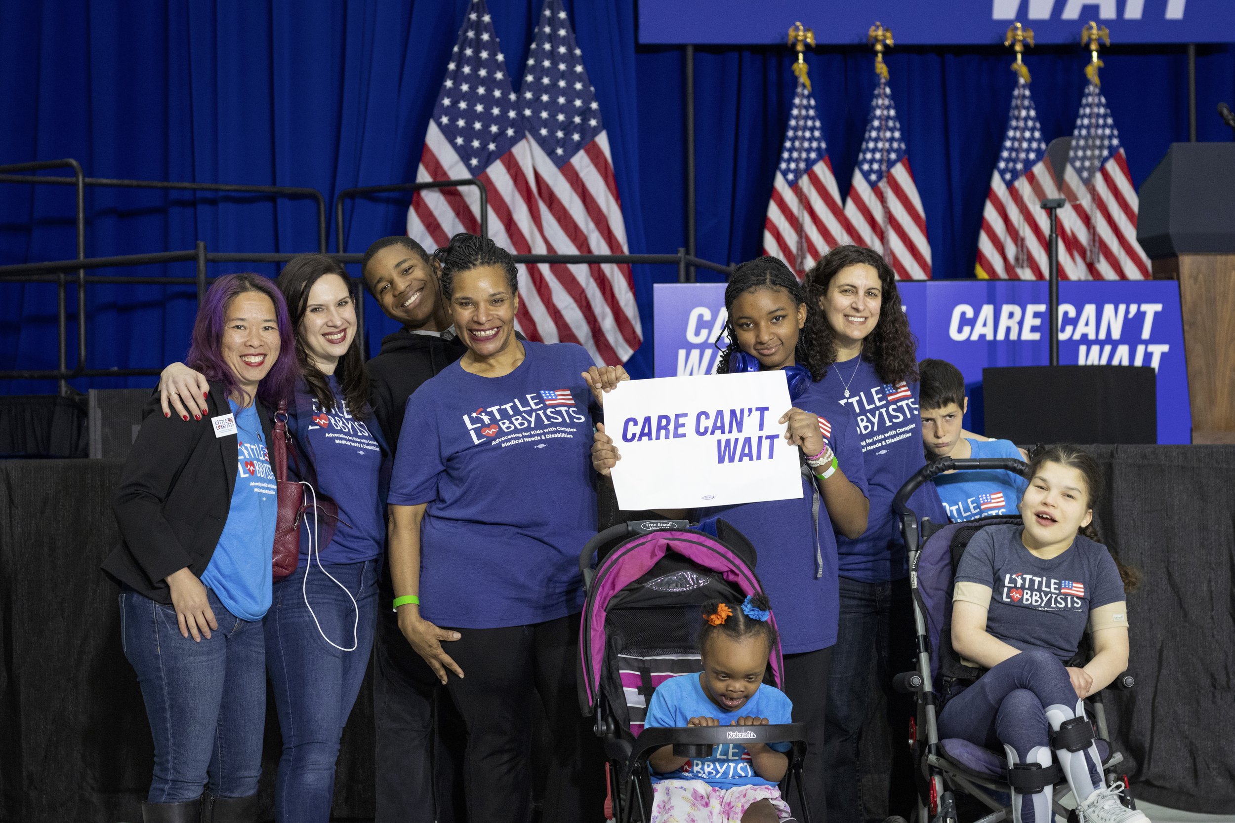  Another group of Little Lobbyists families poses in front of the stage, some standing, some using mobility devices. Danielle’s daughter Autumn holds a sign that says “Care Can’t Wait.” 