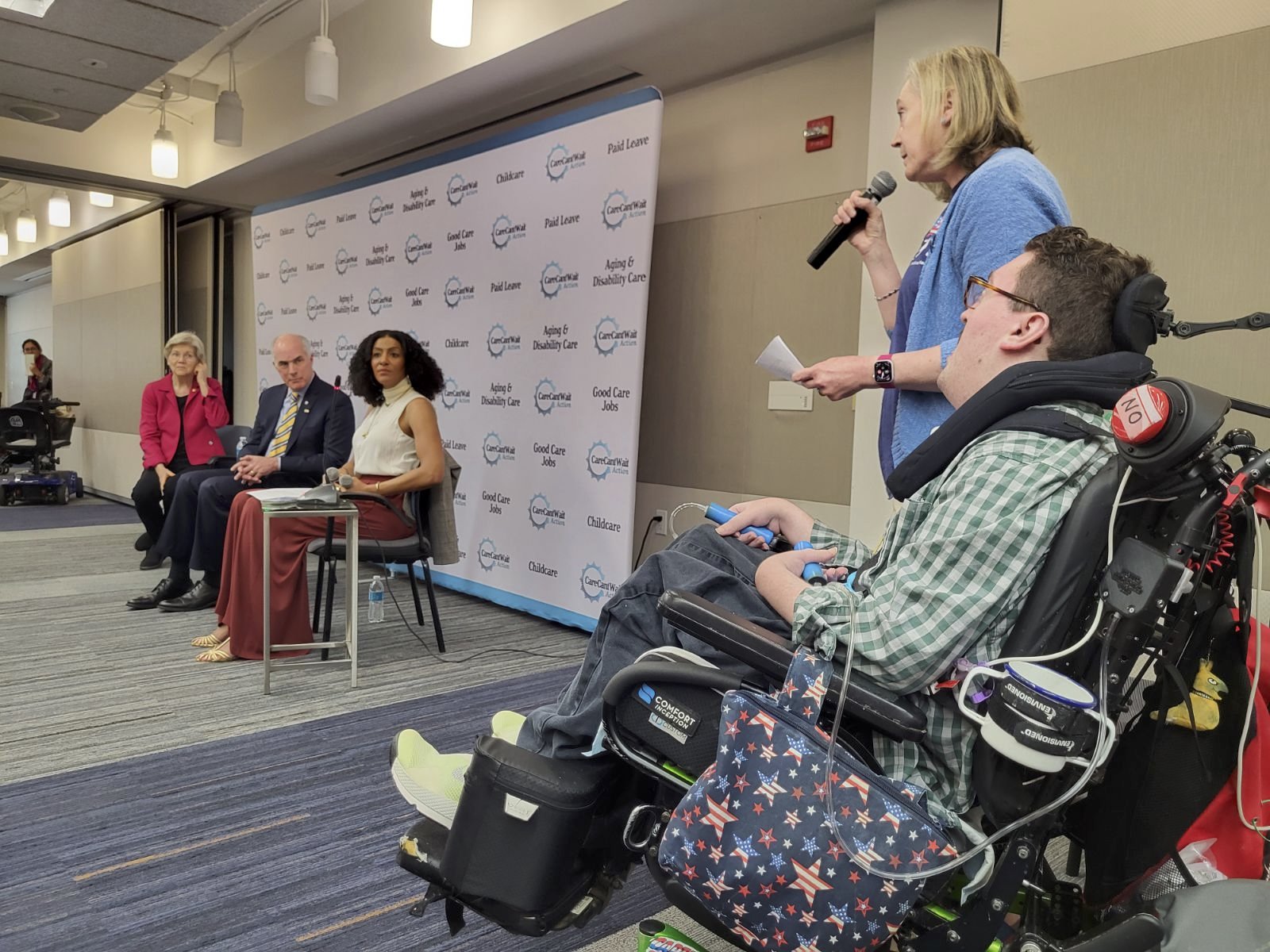  Jeneva holds a microphone, with Rob next to her in his wheelchair, in 3/4 view of their backs. She is asking a question of Senator Elizabeth Warren, who wears a red suit jacket and is seated next to Senator Bob Casey and the host Sarah Jones. 