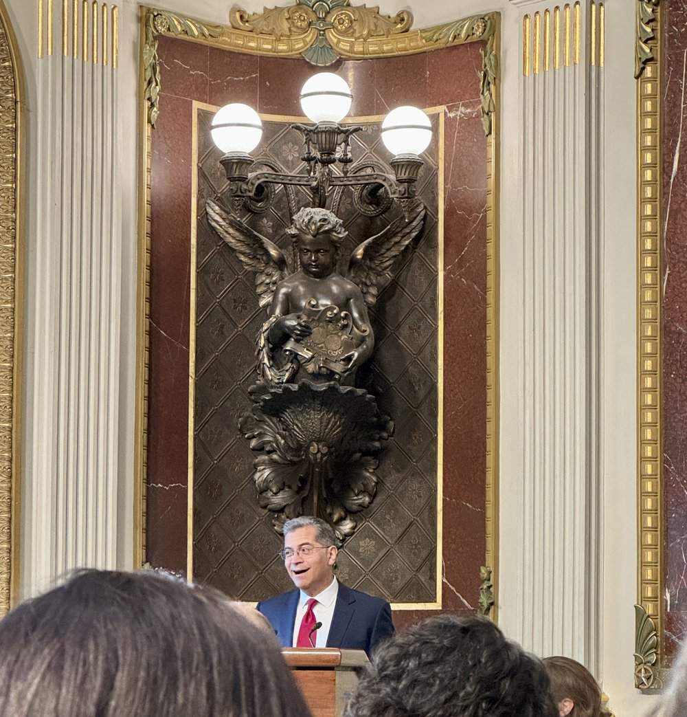  Secretary of Health and Human Services, Xavier Becerra speaks from a podium in an elaborately decorated 19th century meeting room in the Old Executive Office Building, which is adjacent to the White House. Behind him is a large bronze wall decoratio