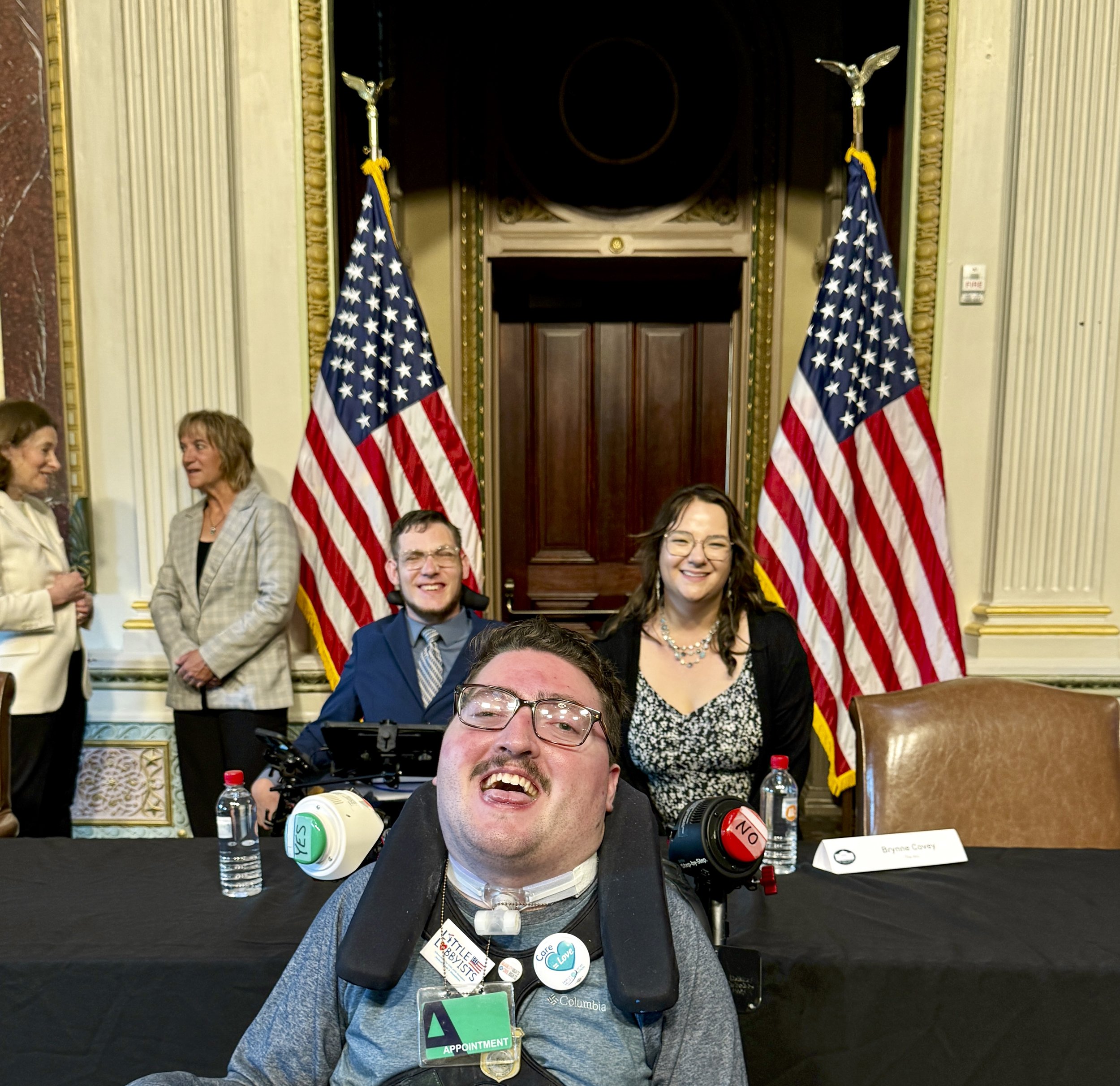  Rob poses in-between advocates Carson Covey (a fellow wheelchair- and AAC-user) and Carson’s sister Brynne. Behind them are two American flags flanking a decorative bronze doorway. 