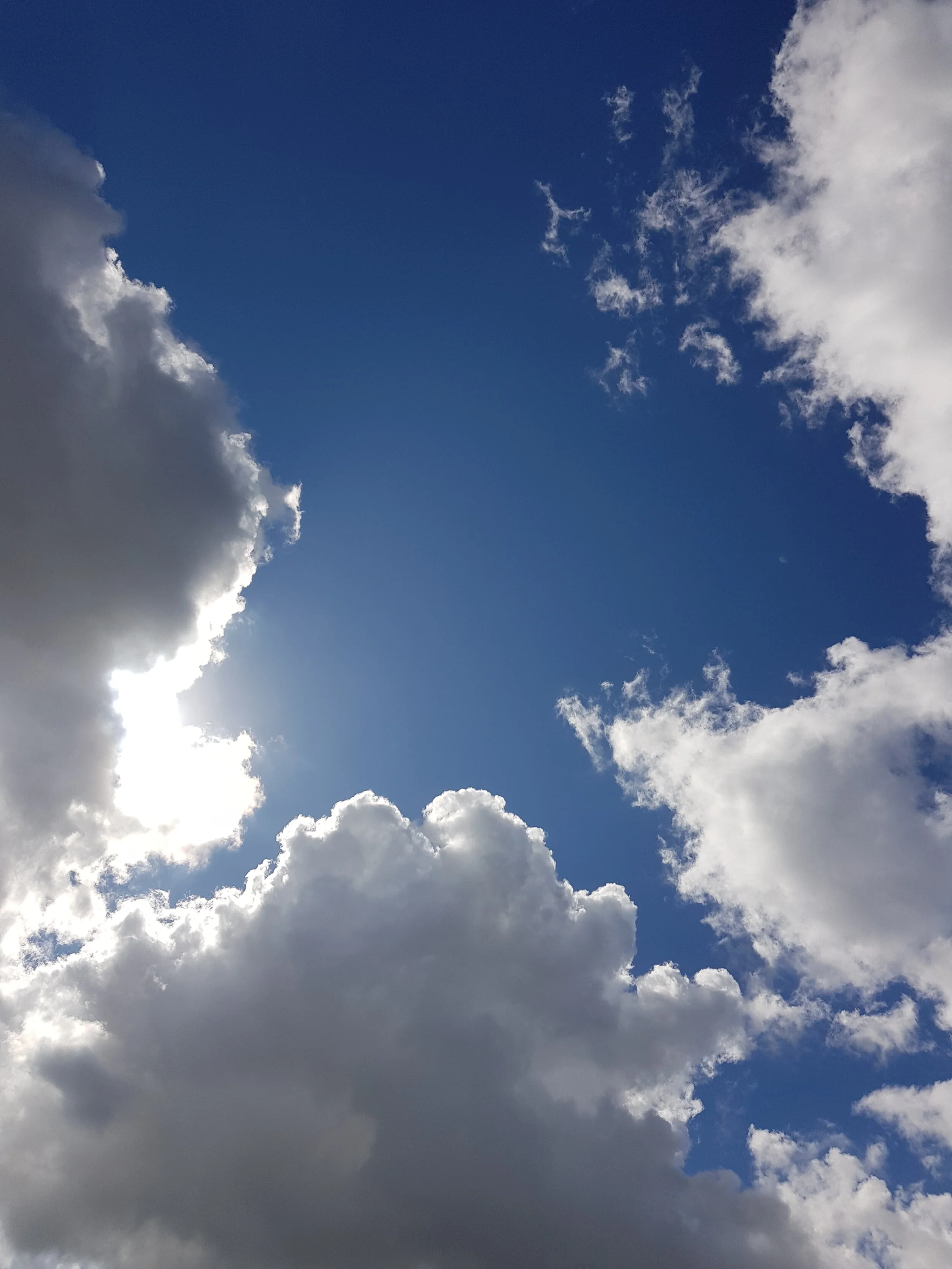 De afbeelding toont een blauwe hemel met enkele grote, witte en grijze wolken, en de zon die schijnt achter de wolken.