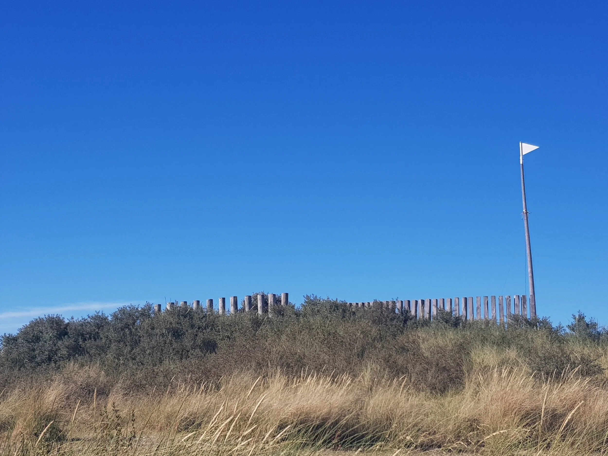Een heuvel met gras en struiken, een houten schutting aan de top, en een hoge vlaggenmast met een kleine witte vlag, onder een heldere blauwe lucht.