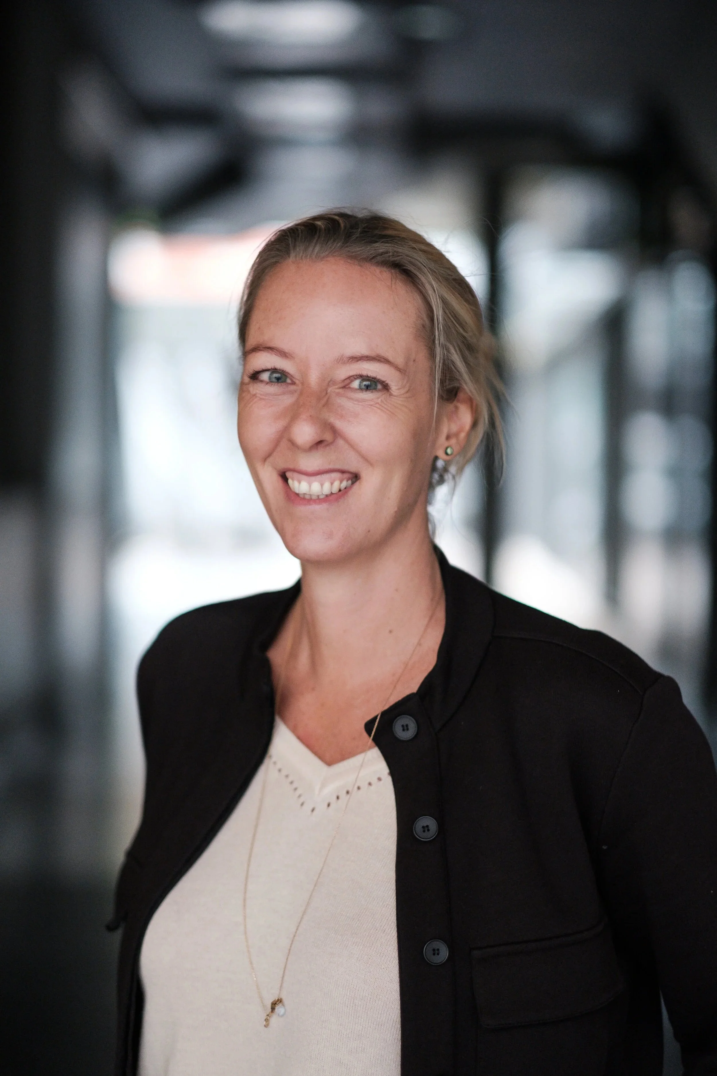 Portret van een lachende vrouw met blond haar, gekleed in een zwarte jas en wit top, in een moderne binnenruimte.