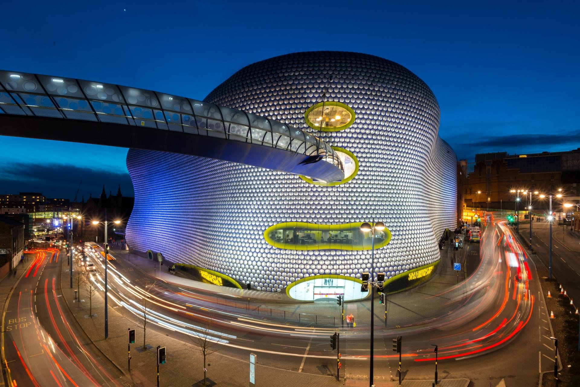 Night view of a modern, bulbous building with circular windows illuminated from within, with a skybridge connecting to another structure, and surrounding streets showing moving cars with light trails.