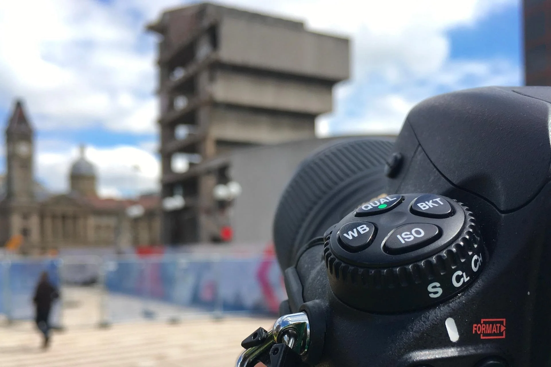 Close-up of a digital camera with control buttons set to a blurred urban background with buildings, a clock tower, and people walking outdoors.