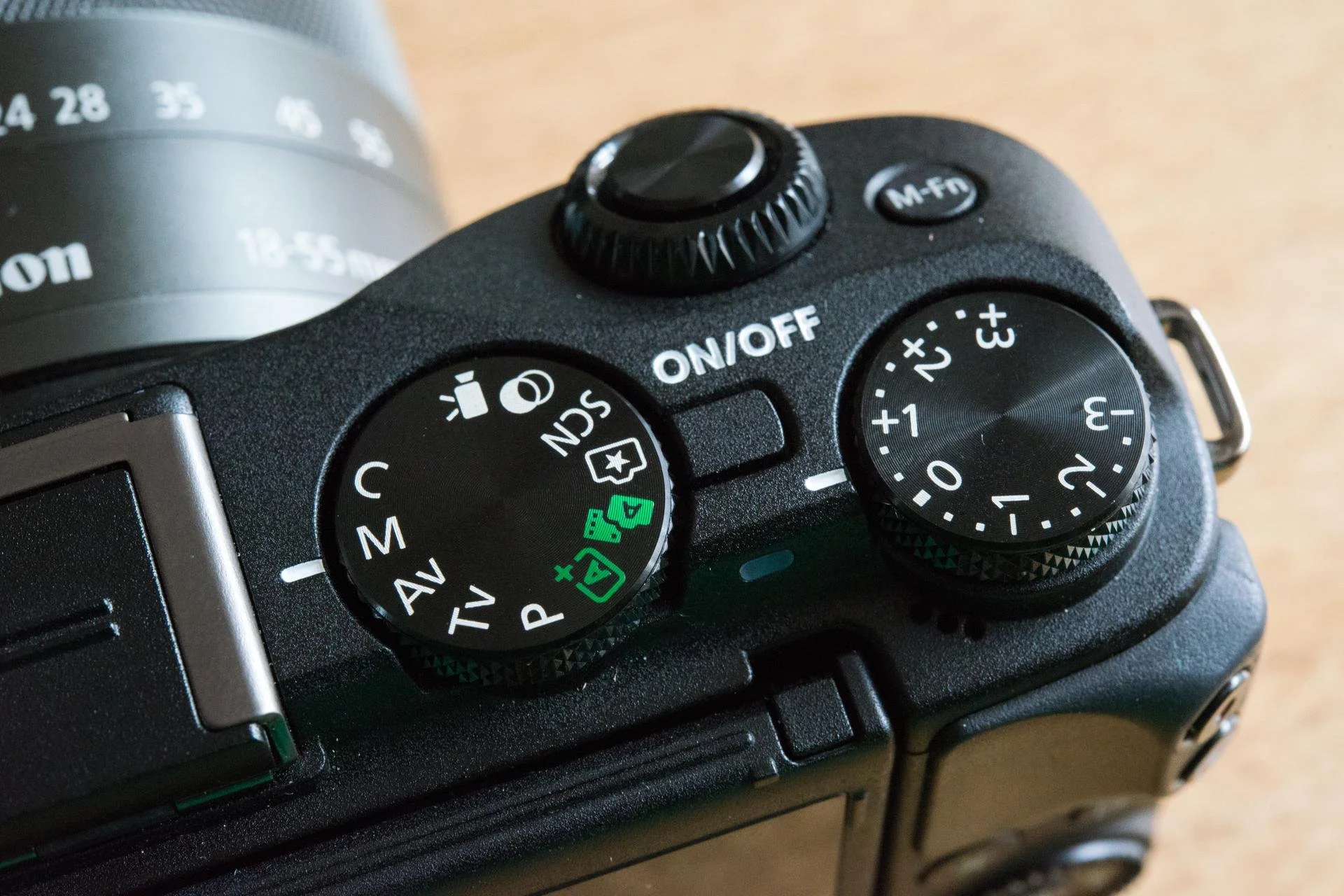 Close-up view of the top controls of a black digital camera, showing two large dials, an ON/OFF switch, and a smaller button marked 'M-FN'.