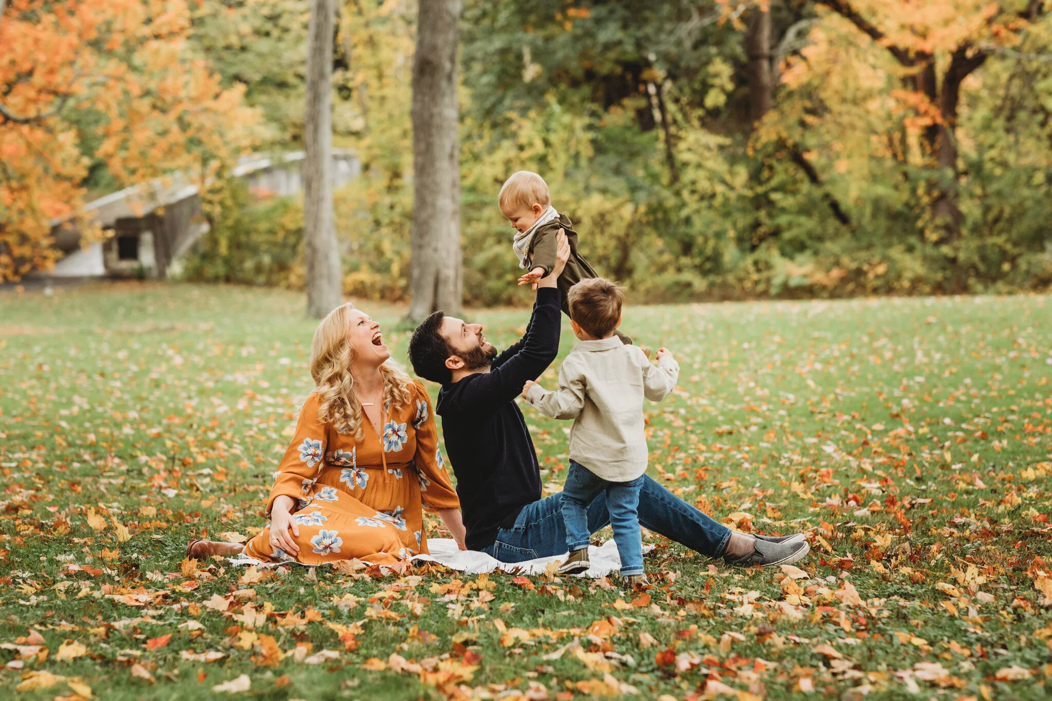 Family of four from Sudbury on a leaf covered lawn with bright foliage behind them during their family photo session. They booked almost a year ahead of time to score this prime foliage spot! Metrowest Boston families: how to score a fall foliage family photography session.