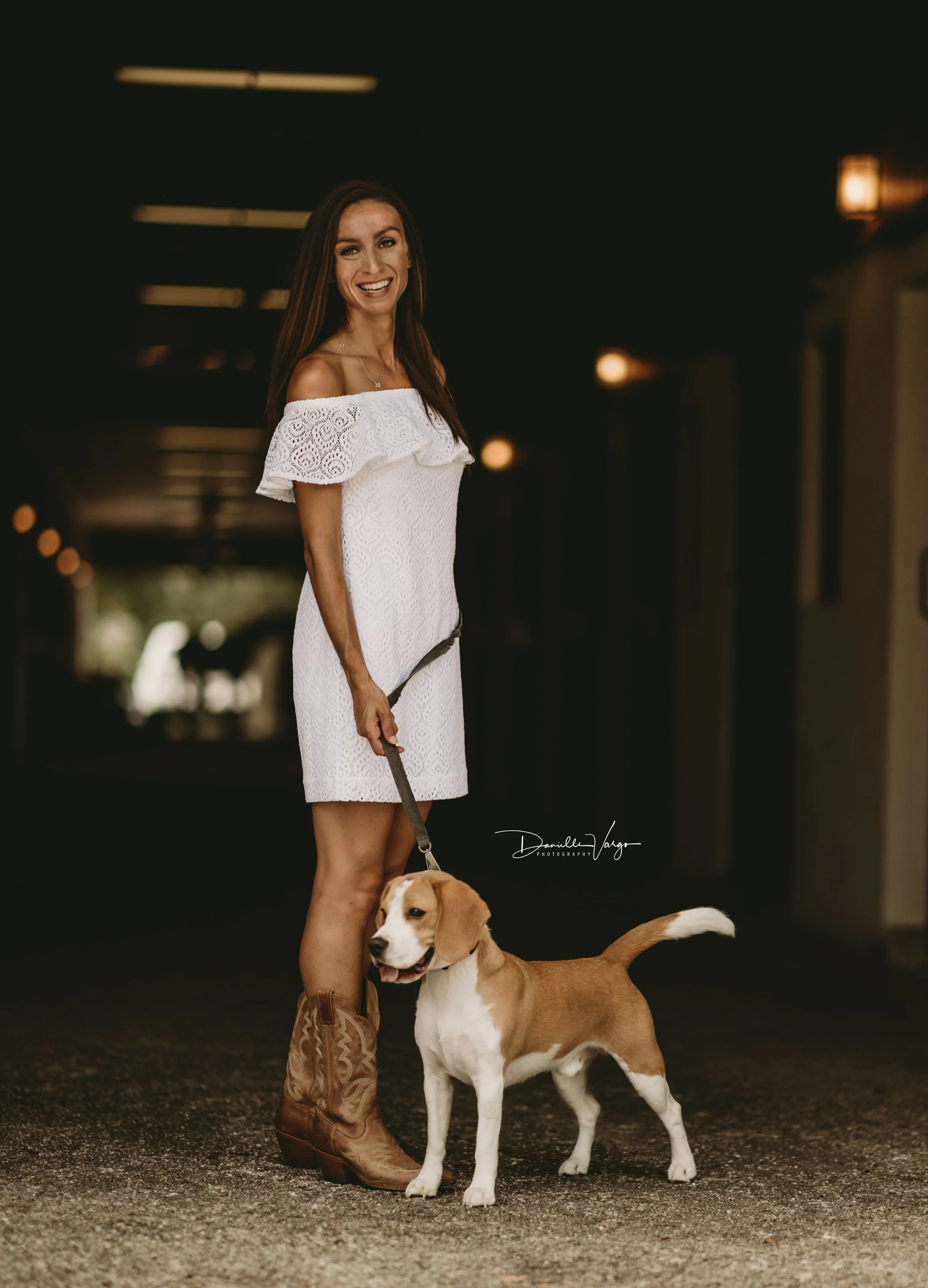 A woman in a white off-the-shoulder dress stands outside under a covered walkway, holding a leash attached to a tan and white dog. She is smiling and looking at the camera, with brown cowboy boots. The background is dark with warm lighting.