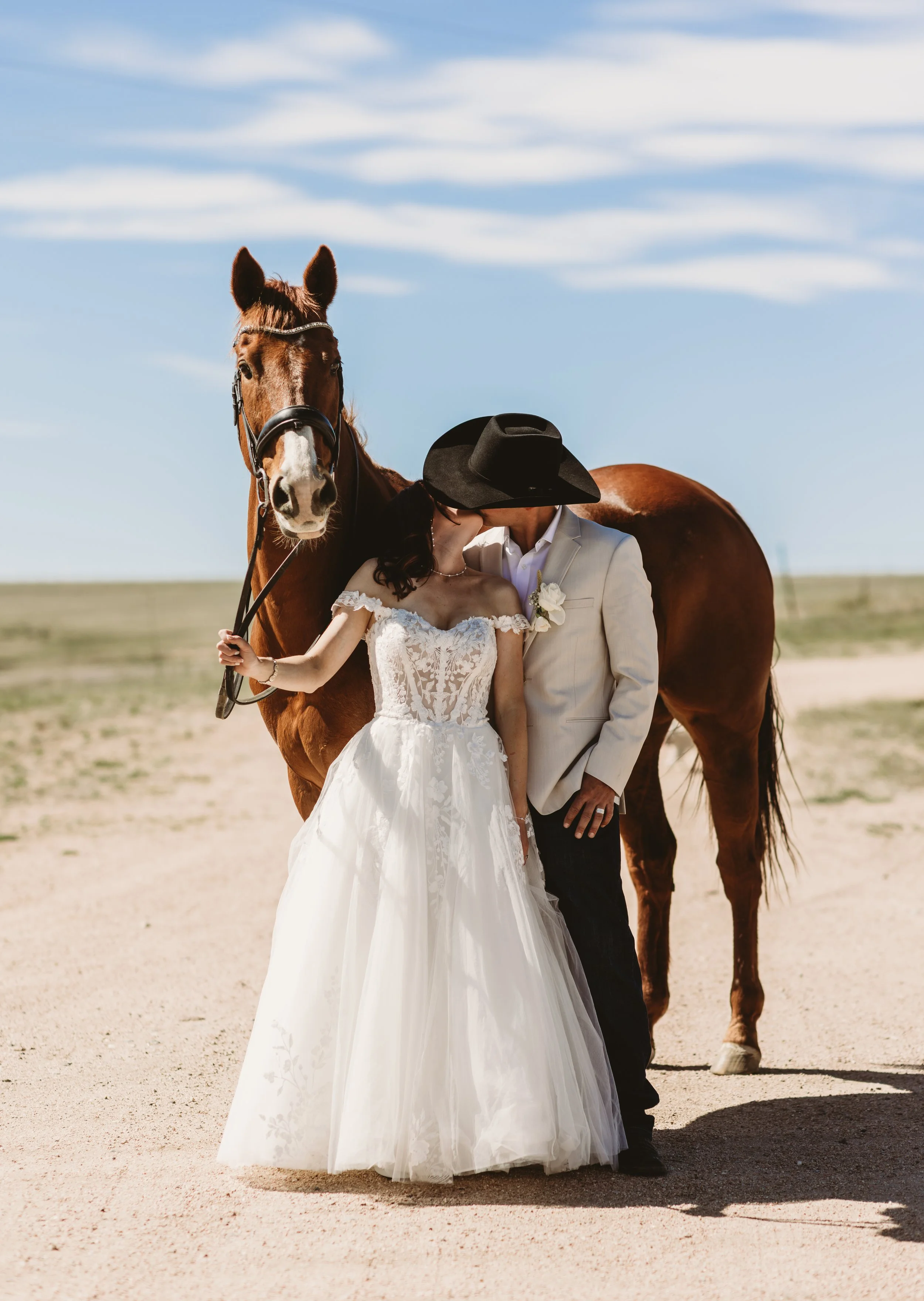 A bride and groom kissing outdoors with a brown horse behind them, under a blue sky with wispy clouds. The bride is wearing a white wedding dress and the groom is dressed in a light-colored suit with a black wide-brimmed hat.