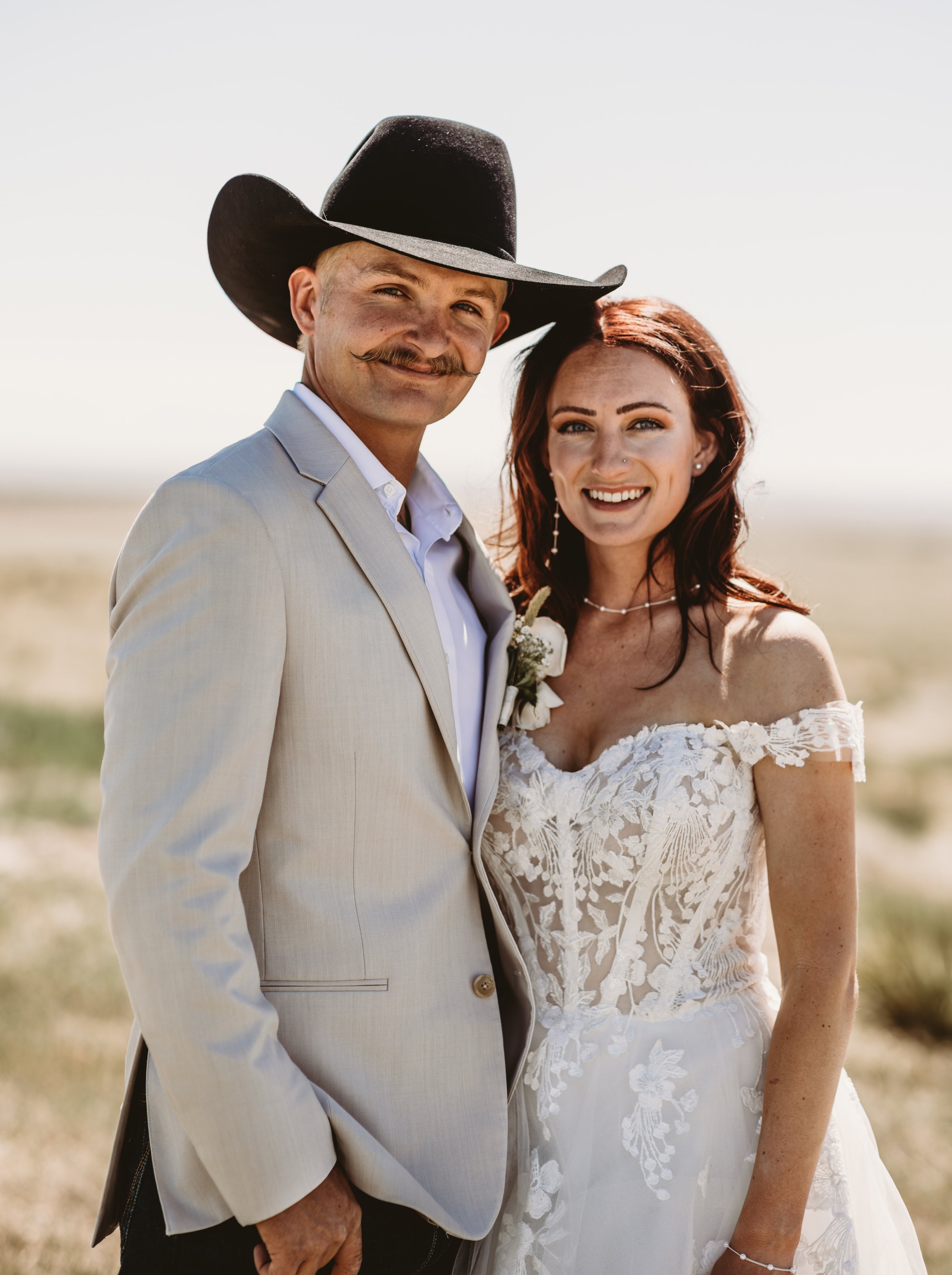 A smiling man and woman dressed in wedding attire standing outdoors on a bright, sunny day. The man is wearing a light-colored suit and a black cowboy hat, while the woman is wearing a white lace wedding dress with off-shoulder sleeves and jewelry.