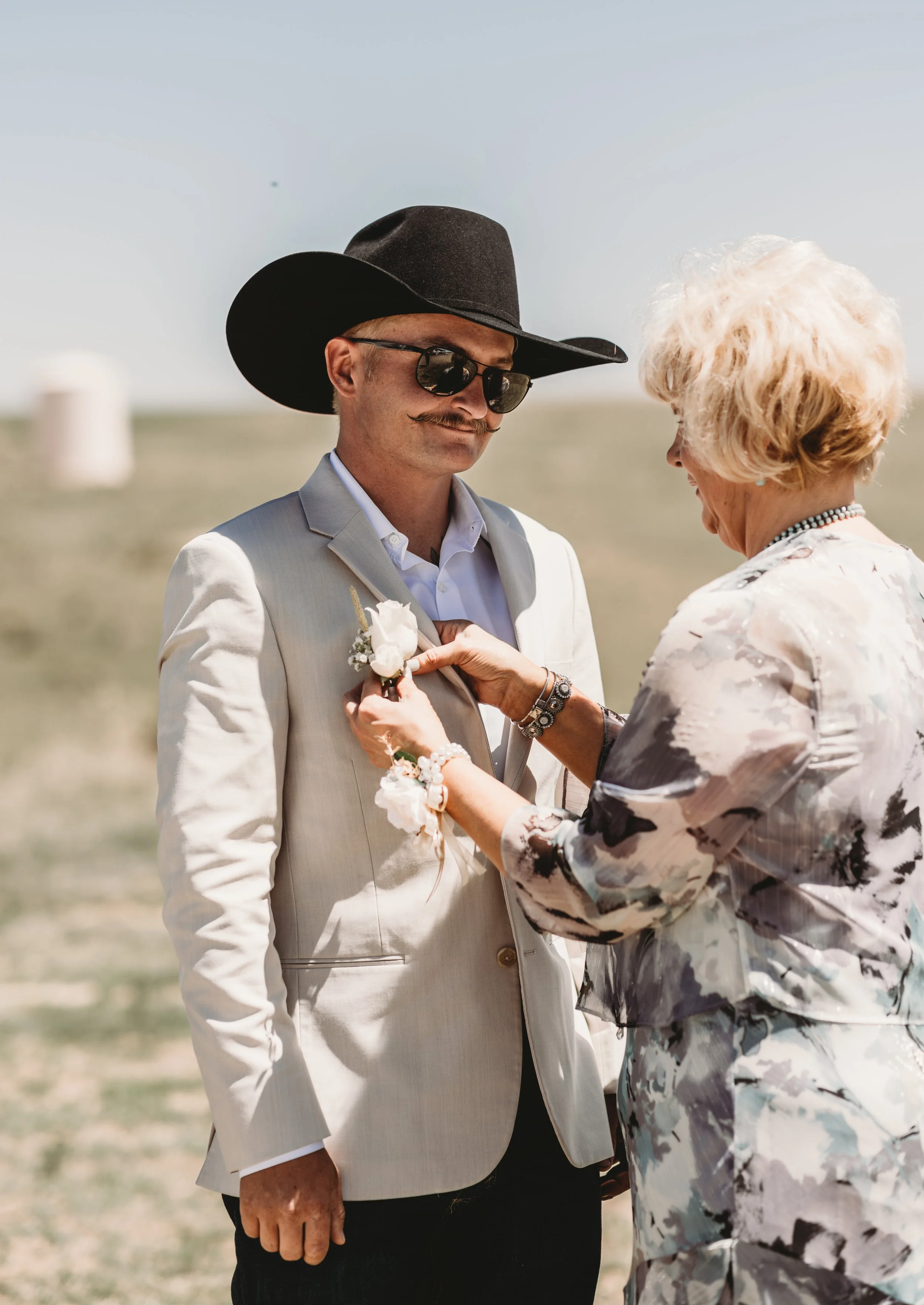 A man wearing a black cowboy hat, sunglasses, and a beige suit with a white shirt, standing outdoors. An older woman with blonde hair, wearing a floral-patterned dress, is pinning a boutonniere on his suit. They are smiling at each other with a clear