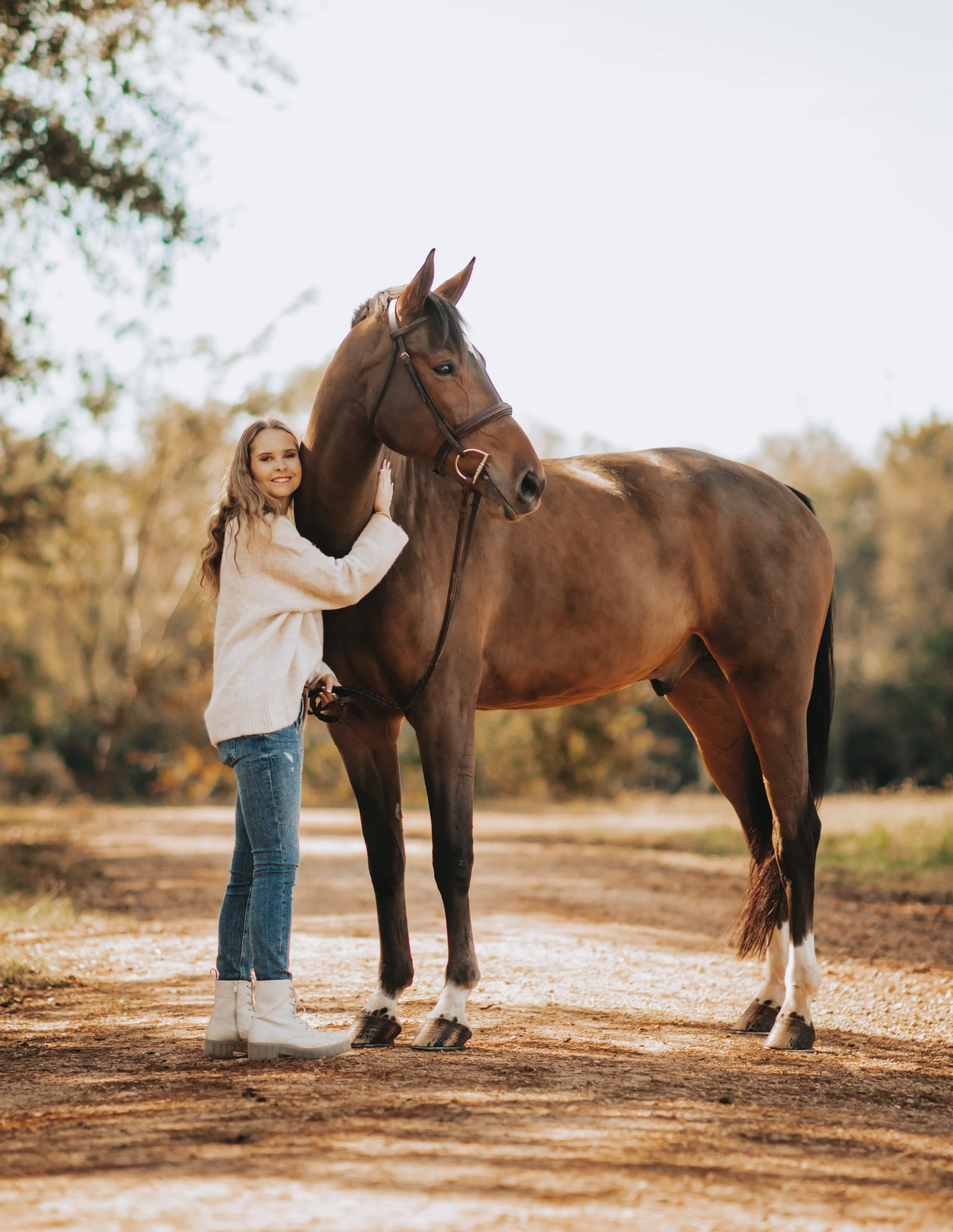 A young woman with long wavy hair, wearing a cream sweater, jeans, and white boots, stands next to a brown horse with a white star on its forehead, on a dirt path surrounded by blurred autumn trees.