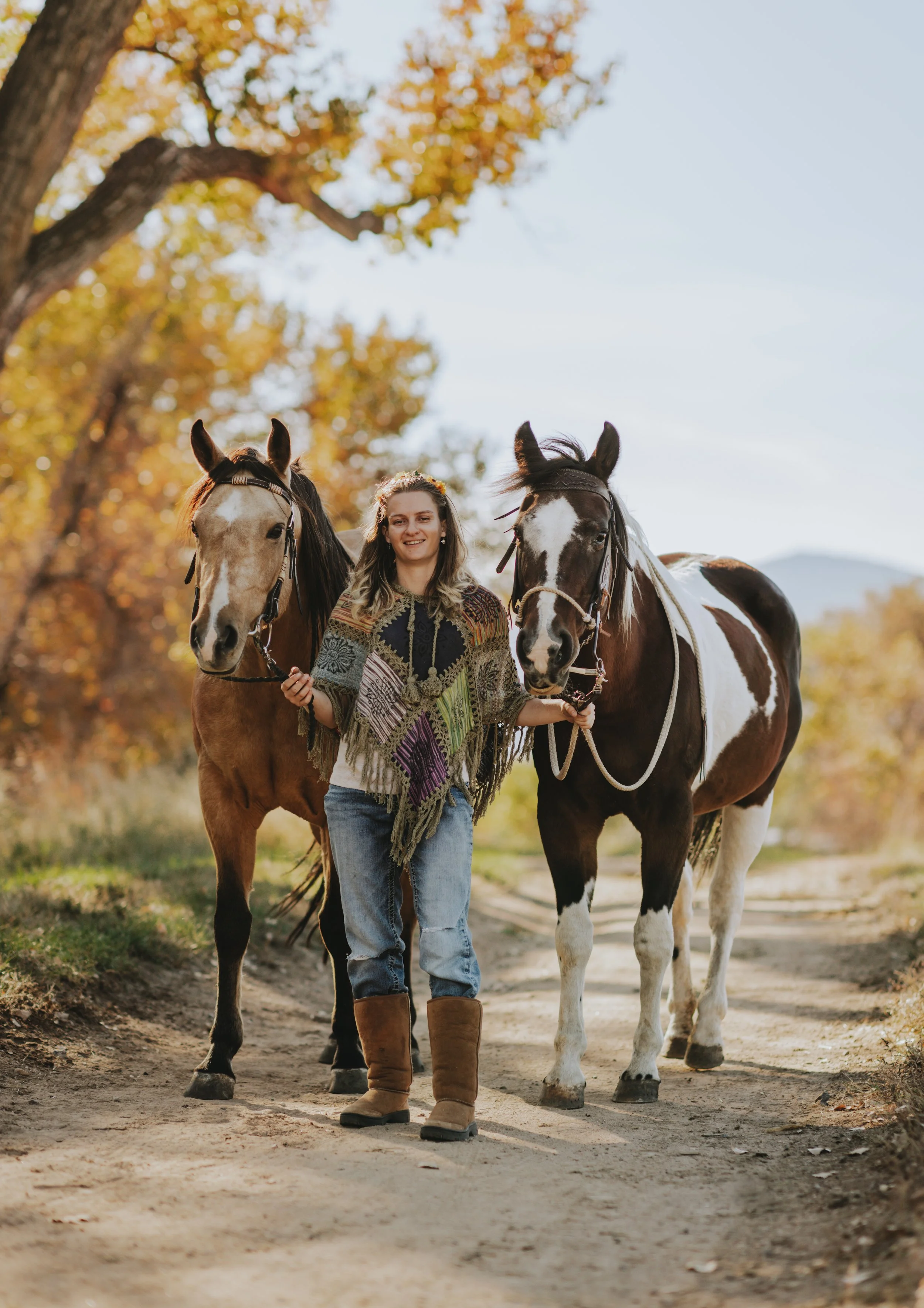 A woman walking outdoors with two horses during autumn, holding the reins of each horse, with trees in the background and a clear sky.